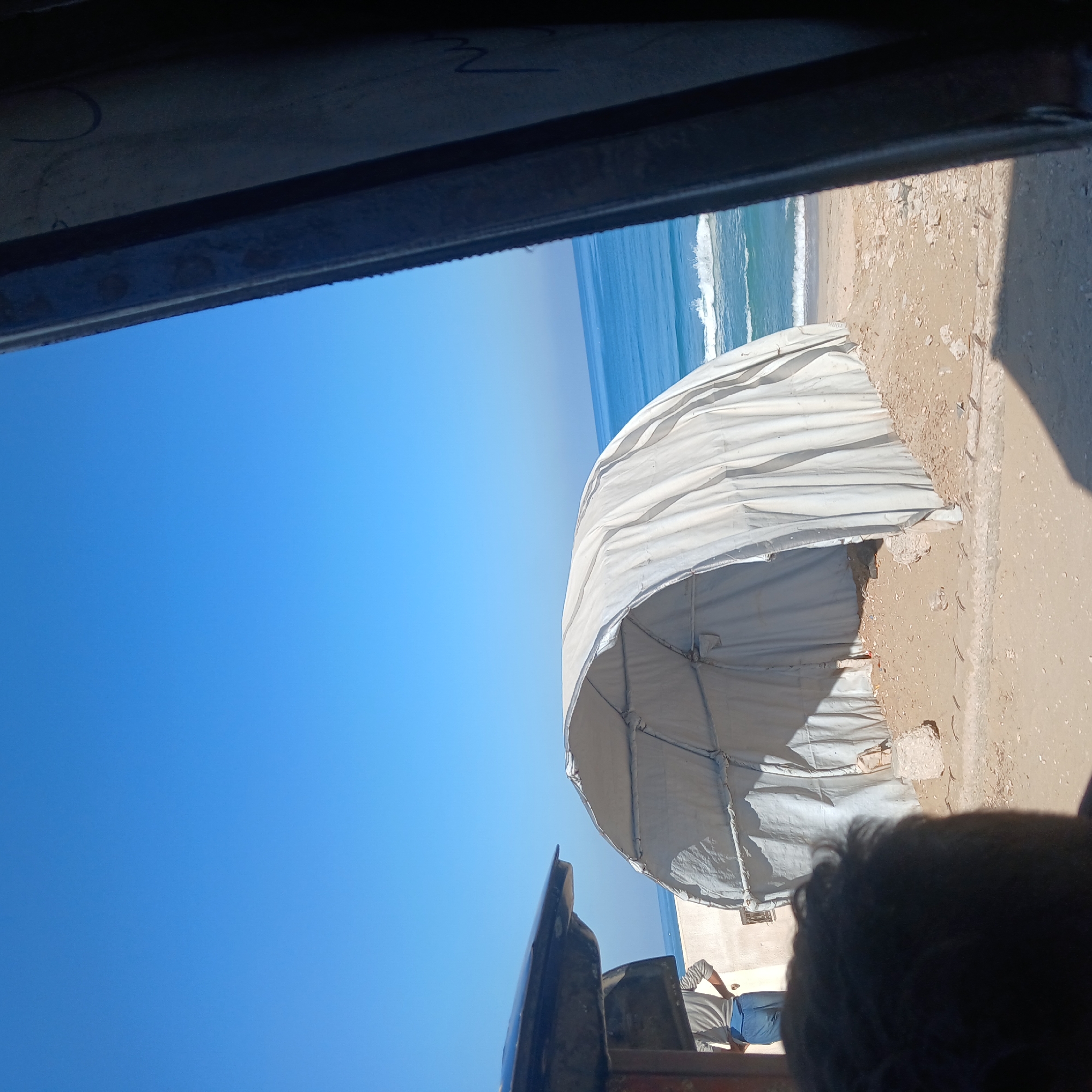 The image depicts a beach scene through a frame, possibly a window or vehicle. The sky is bright blue, transitioning to a horizon with a visible coastline and ocean waves. On the right side of the frame, a white, dome-shaped structure resembling a tent or canopy sits on the sand. The sand is light brown and appears wet in places. A portion of a building or other structure is visible behind the dome. In the foreground, at the bottom right, a dark-haired individual is partially visible. The upper left and lower areas of the frame are dark, indicating the edges of the viewing structure. The location is likely a coastal area in an unknown city.