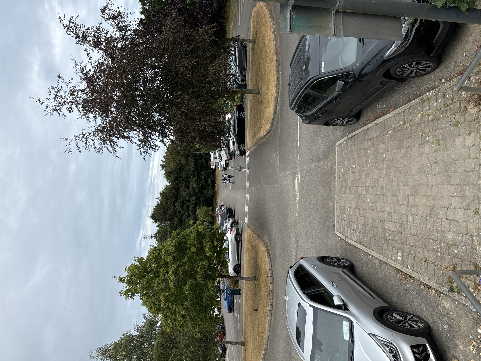 The image shows an outdoor parking lot with several cars parked. In the foreground, a silver SUV is parked on the left, and a black SUV is parked on the right, partially obscuring a utility pole. In the middle ground, a row of cars is parked along the side of the lot, including a blue sedan and several silver and black vehicles. Several people are walking across the parking lot in the distance. The parking lot is paved, with grass and trees surrounding it. The sky is overcast with clouds. The location is identified as Courtown, Ireland.