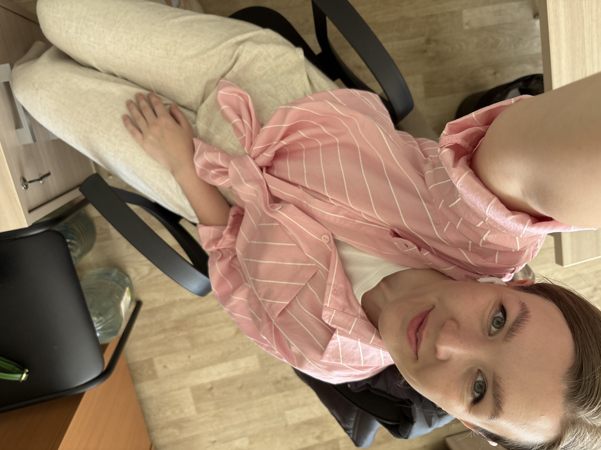 A high-angle, close-up selfie shows a fair-skinned woman sitting in a black office chair. She has brown hair pulled back, green eyes, and is wearing a white shirt underneath a pink and white striped button-down shirt tied at the waist. She is also wearing light-colored pants. The woman is looking up at the camera. A desk or cabinet is visible to the left of the chair and a light-colored wooden floor is in the background. The city of Izhevsk, Russia is not discernable from the image.
