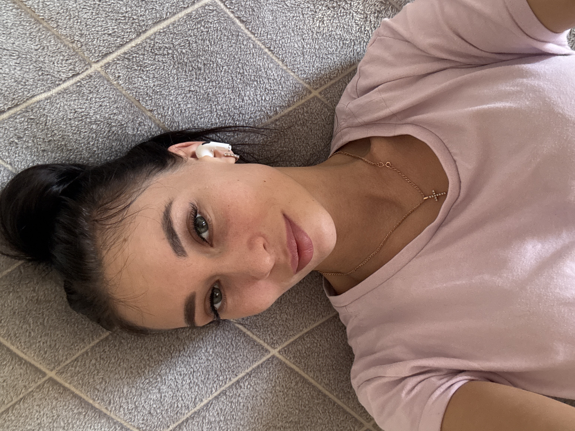 A young woman with dark hair pulled back in a bun is lying on a gray tiled floor. She is wearing a light pink t-shirt and a gold necklace with a cross pendant. She is looking at the camera. There are white earbuds in her left ear. The location is unknown, and it is not possible to determine if this image was taken in Chaikovsky, Russia.