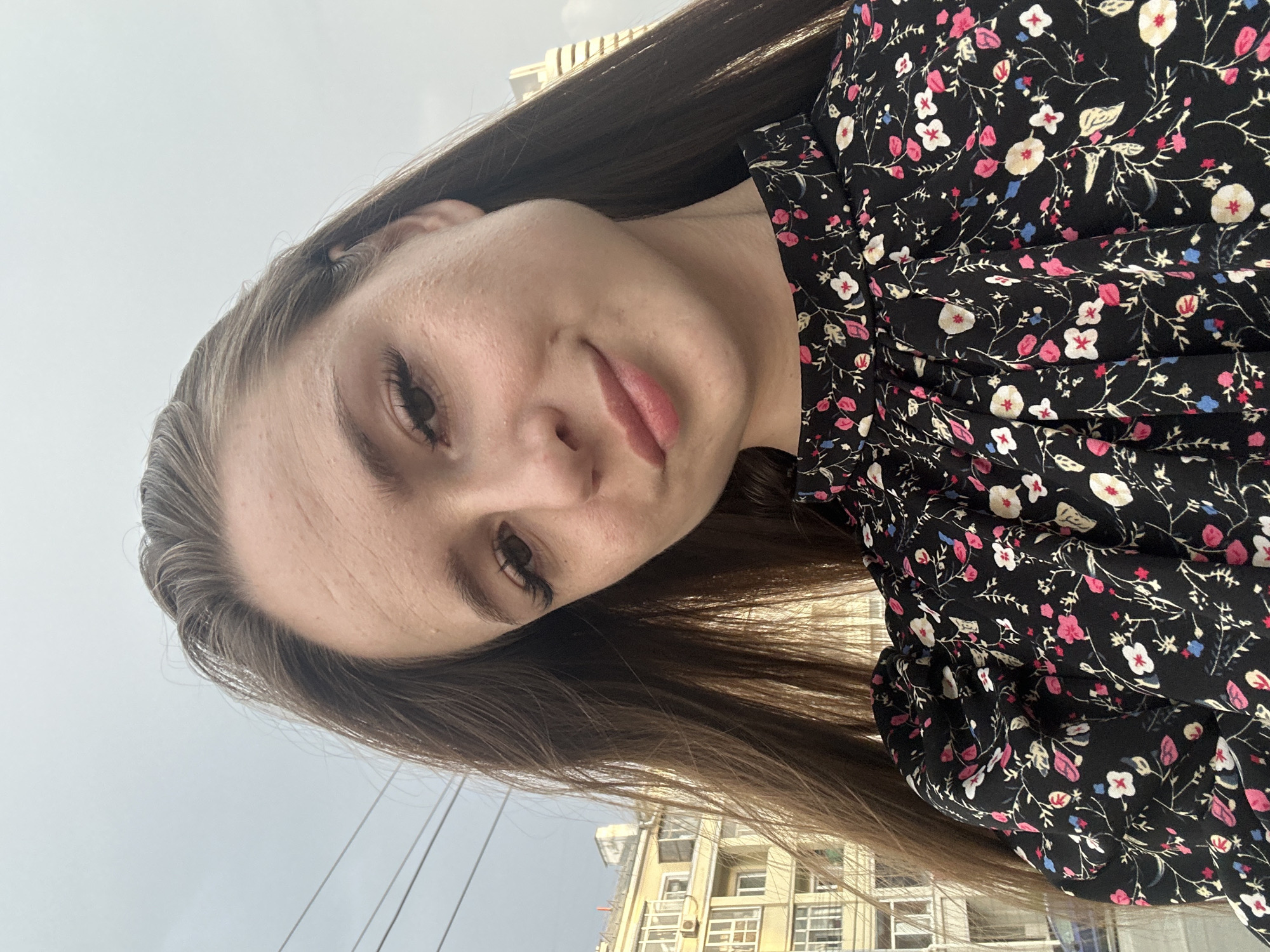 A woman with long brown hair is shown in a close-up, outdoor selfie. She is wearing a black dress with a floral pattern. Her face is the primary focus of the image, with her hair framing her face. In the background, out of focus, are buildings and utility wires, suggesting an urban environment. The sky is overcast.