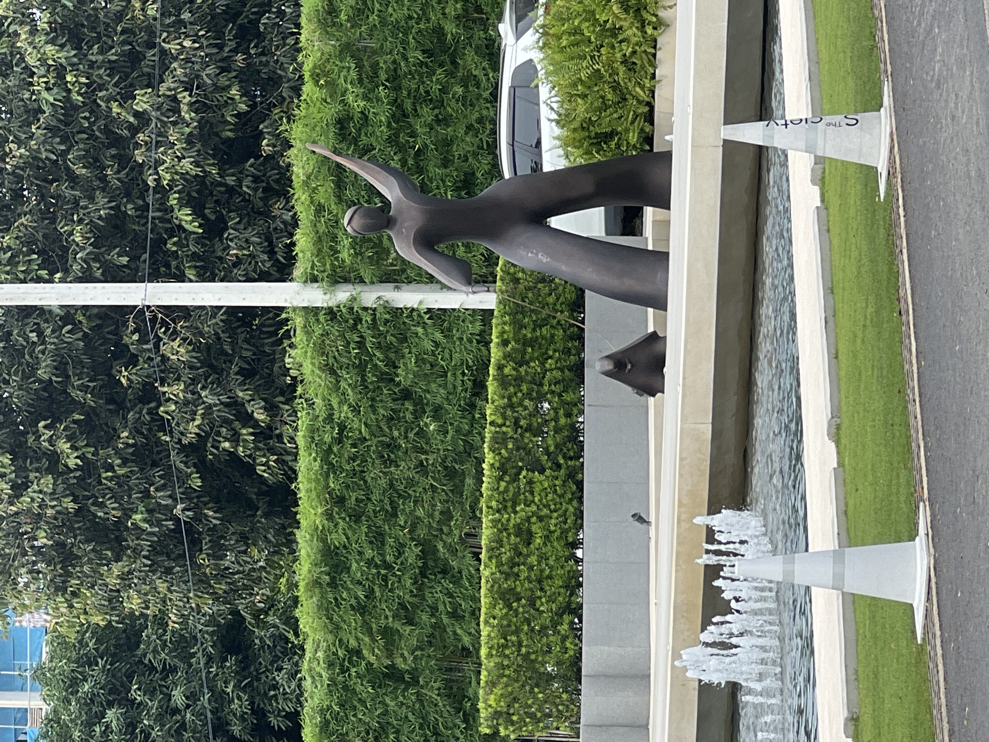 The image depicts a statue of a person with arms outstretched, positioned near a fountain. The statue appears to be made of bronze or a similar dark metal. The fountain has multiple spouts of water. The statue and fountain are located near a roadway, as indicated by the presence of white traffic cones. Behind the statue and fountain is dense greenery, including trees and bushes. In the background, a building is partially visible. The location appears to be in an urban setting with landscaping. The image is taken from a ground-level perspective.