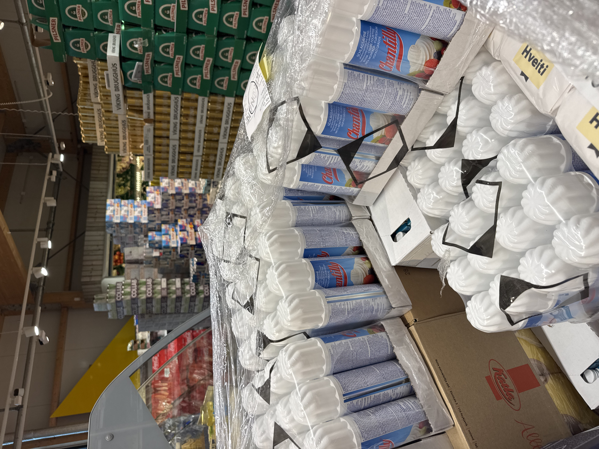 The image shows a stack of products in a store, presumably in Vik, Iceland, based on the visible text "Katla matvælaiðja ehf. Kletthåls 3 110 Reykjavík Iceland Tel" on one of the boxes. There are rows of "Pilsner" and "Viking Brugghús" beer boxes in the background. Closer to the camera, there is a stack of "Chantilly" whipped cream cans, bags labeled "Hveiti" (Icelandic for flour), and boxes with a logo of "Katla," a food producer. The items are mostly packed in plastic wrap.