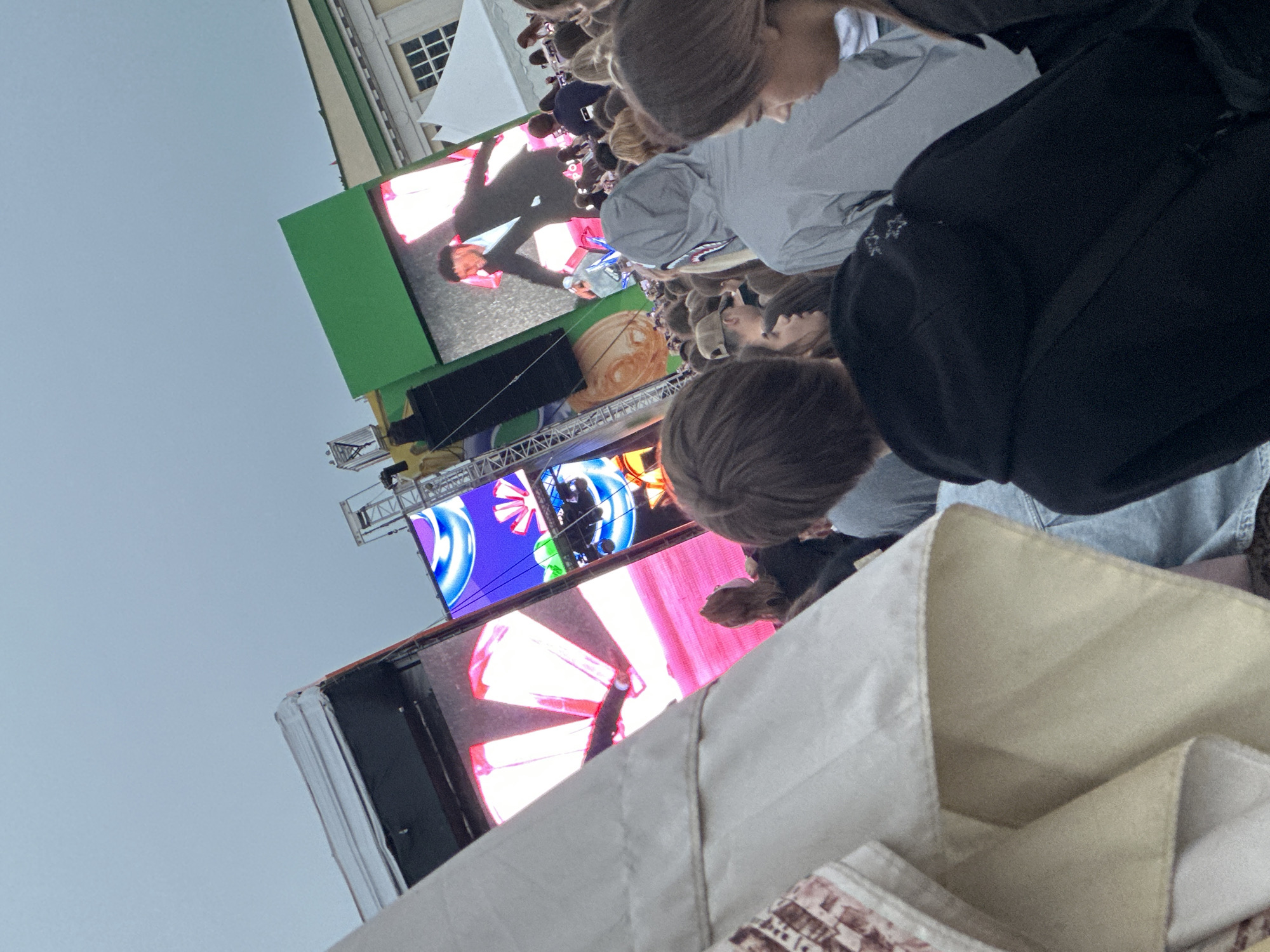 The image captures an outdoor event in Minsk, Belarus, under an overcast sky. A large crowd of people, mostly young adults, is visible from behind and in profile, facing a performance stage.
In the immediate foreground, a significant portion of a light-colored fabric canopy or tent, with visible folds and seams, extends from the left, partially obscuring the lower-left corner of the scene.
The central area is dominated by a complex stage structure featuring multiple large LED video screens supported by metal trussing. The screen on the far right displays a male performer in a dark suit and light shirt, captured in a dynamic pose with an outstretched arm. Other screens exhibit bright, abstract digital graphics, including swirling blue and white patterns, a partial NBC-style circular logo, and prominent angular shapes in pink and red tones. A person dressed in yellow is visible on a ladder-like structure adjacent to the central screens, appearing to work on the stage setup.
The crowd, filling the mid-ground, is diverse in appearance, with individuals wearing casual clothing such as hoodies and baseball caps. One person in the foreground wears a black hoodie with light-colored detailing on the back of the hood. Another person in the mid-ground wears a grey hoodie with a distinctive shark-face graphic on the hood. The crowd is uniformly oriented towards the stage.
In the background, a classical-style building with a light-colored facade, green roof elements, and white-framed windows is visible behind the stage. A white event tent is also present to the right of this building. The sky above is a uniform light grey, indicating an overcast day.