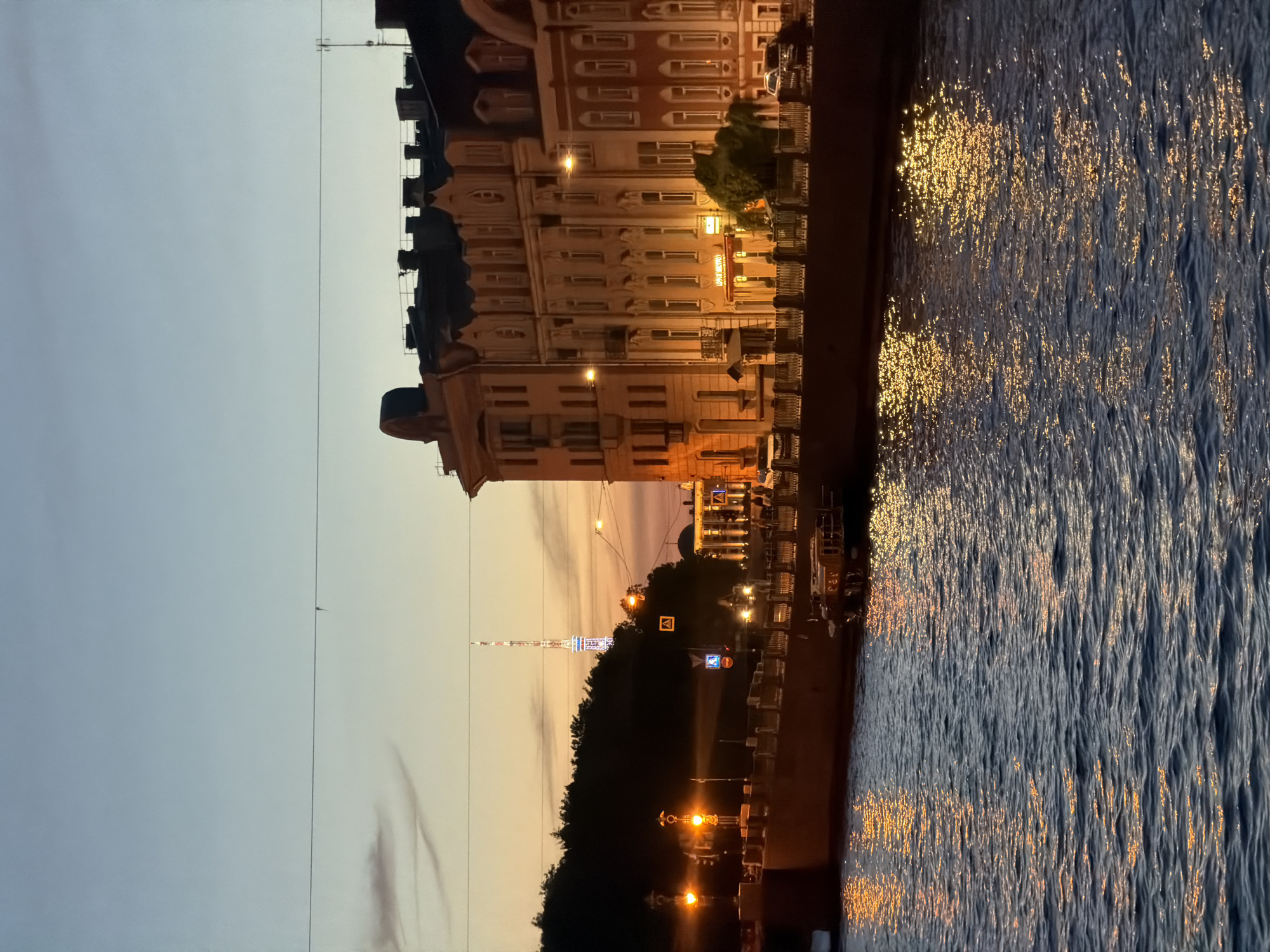 The image depicts a twilight view of a canal in Saint Petersburg, Russia. On the right bank, a row of historic buildings with ornate facades and mansard roofs line the canal. The buildings are illuminated by interior lights and street lamps. A prominent television tower is visible in the distance against the darkening sky, which transitions from a pale orange at the horizon to a soft blue overhead. The canal water reflects the lights from the buildings and street lamps, creating shimmering patterns. A small boat is moored along the embankment. The scene suggests an evening in the city, with a mix of historic architecture and modern infrastructure.