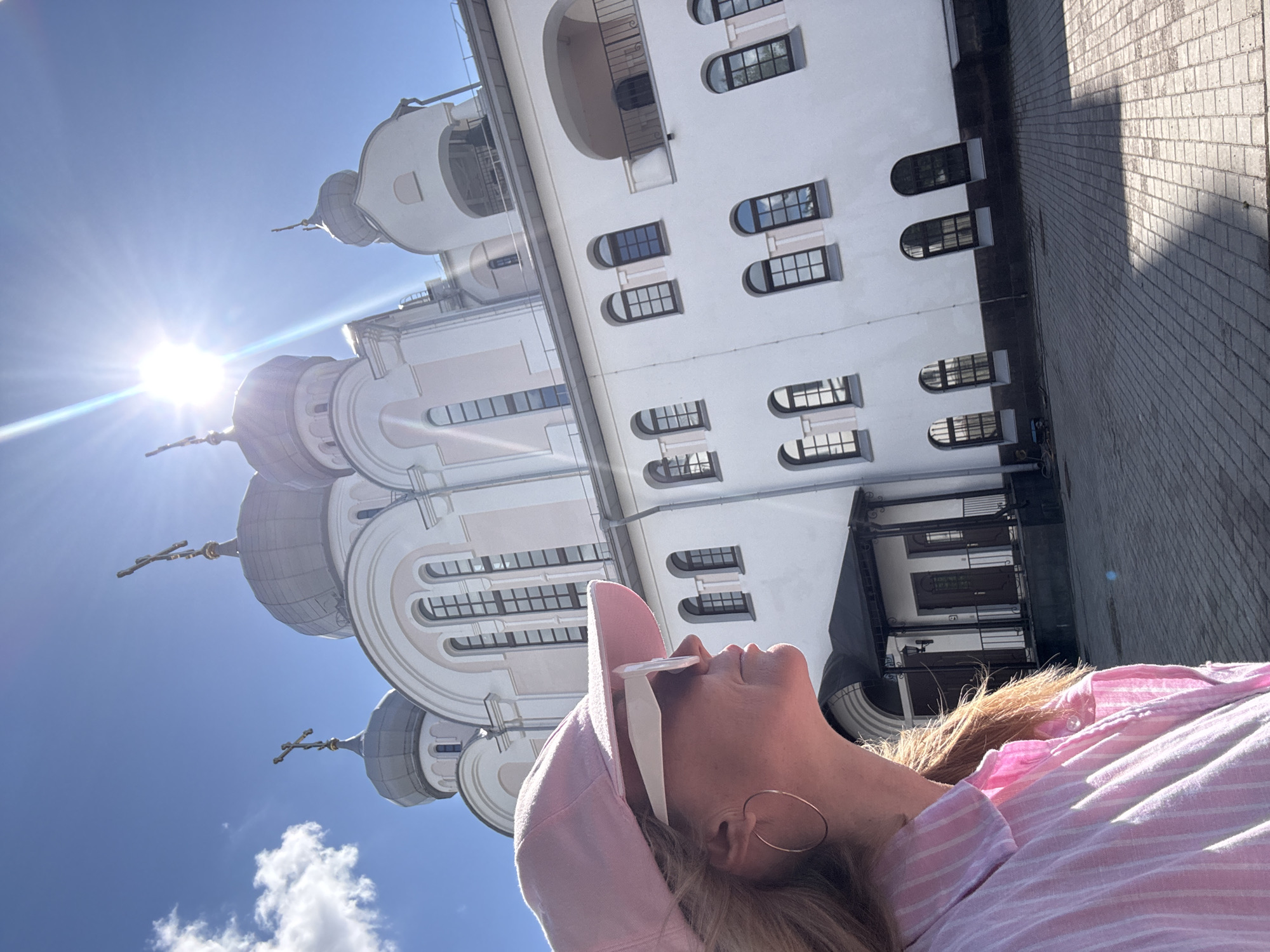 The image shows a woman in a pink striped shirt and pink baseball cap, wearing large white sunglasses and gold hoop earrings. She is looking up and to the right, with her profile visible. Behind her, a white building with multiple domes and crosses stands against a bright blue sky with a few clouds. The sun is shining intensely, creating lens flares. The building appears to be a church or monastery. The ground in front of the building is paved with cobblestones. The architectural style of the building, particularly the domes and crosses, suggests a Russian Orthodox church. Given the style of the architecture and the bright, clear sky, this scene is likely located in Saint Petersburg, Russia, a city known for its numerous churches and cathedrals.