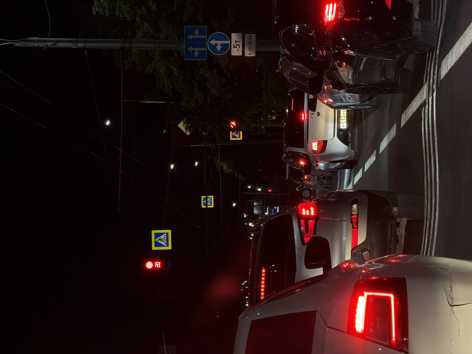 This image captures a nighttime traffic jam in an urban environment. Several cars are visible, mostly from the rear, with their brake lights illuminated, indicating they are stopped or moving slowly. The foreground features the rear of a white car. Behind it are several other vehicles, including a silver SUV and a white hatchback. Further down the road, more cars are seen.
Various traffic signs and signals are present. A traffic light shows a red circular signal with a "20" displayed below it, suggesting a speed limit. Yellow triangular signs with blue pedestrian icons indicate a crosswalk. A blue square sign with white arrows indicates lane directions. Another blue circular sign with a white arrow and "5T" below it likely refers to a weight restriction, with "6.00-21.00" indicating the time of day it is in effect. Overhead wires, possibly for public transportation, are also visible. Streetlights cast illumination on the scene, and trees can be seen on the right side of the image. The overall atmosphere is one of a congested city at night.