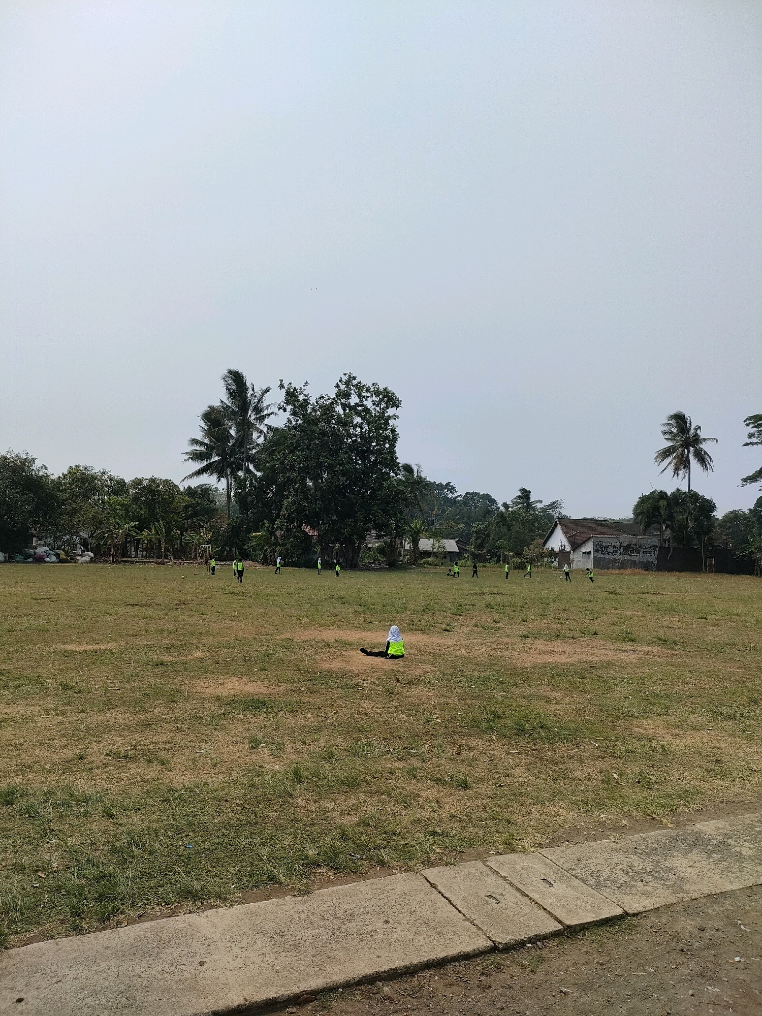 The image depicts a wide, open field in what appears to be a rural or semi-rural area, likely Bojong, Indonesia. The field is covered in dry, yellowish-brown grass with patches of greener vegetation, suggesting a dry season or drought conditions. In the background, a line of lush green trees and vegetation forms a natural border, with several buildings and homes interspersed among them. Two palm trees are prominent in the middle ground, one tall and slender, the other shorter and fuller. A soccer goal is visible in the left-center of the field. Several individuals, dressed in bright green shirts, are scattered across the field, seemingly engaged in a soccer game. One person, wearing a bright green hijab and shirt, is seated on the grass in the foreground, facing away from the viewer. The sky is overcast and pale, with no visible clouds, indicating a diffused light. In the immediate foreground, a concrete path or sidewalk is visible, with drainage covers embedded in it. The overall impression is a community space utilized for recreational activities.