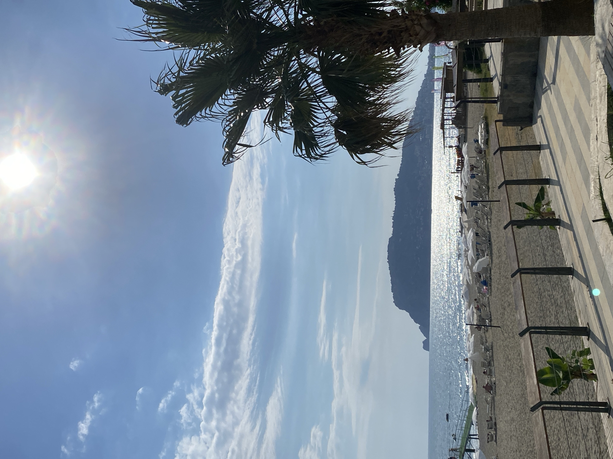 The image depicts a scenic beach scene in Alanya, Turkey, on a bright, sunny day. The sun is high in the sky, casting a strong glare on the water, suggesting it is midday. The weather appears to be warm and clear, ideal for a beach outing.
The foreground is dominated by a palm tree, its fronds casting shadows and framing the top left portion of the image. To the right, a paved walkway with a tiled pattern runs alongside the beach. This walkway is lined with a modern metal railing.
The beach itself is sandy and appears to be well-equipped with numerous white beach umbrellas and loungers, indicating a popular tourist spot. Some people are visible on the beach, relaxing under the umbrellas or near the water. The sea sparkles under the sun, stretching out to the horizon where a dark, mountainous coastline is visible. The calm water suggests a tranquil atmosphere.
In the distance, a pier or jetty extends into the sea, with some people gathered on it. The overall mood of the image is one of relaxation and enjoyment of a beautiful summer day at the seaside. No visible text is present in the image.
