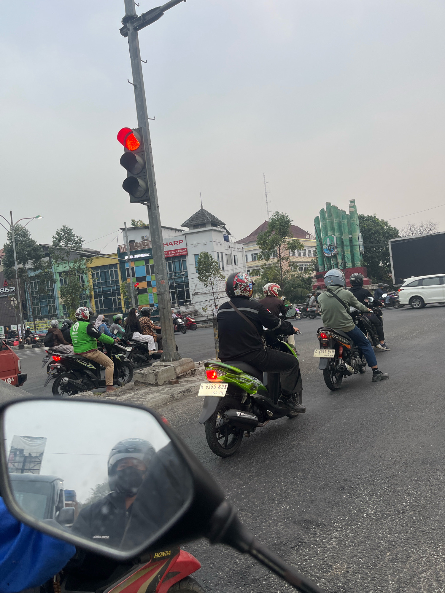 The image depicts a street scene in Bekasi, Indonesia, likely near a busy intersection. A traffic light is visible, showing a red signal, indicating that vehicles should stop. Several motorcycles are present, with riders wearing helmets, some of whom appear to be wearing masks. One motorcycle has a green Gojek jacket, suggesting a ride-hailing service. In the background, there are buildings, including one with a "SHARP" sign. A distinctive green monument, possibly a landmark in Bekasi, is also visible. The sky is overcast, suggesting a cloudy day. The perspective suggests the photo is taken from another motorcycle.