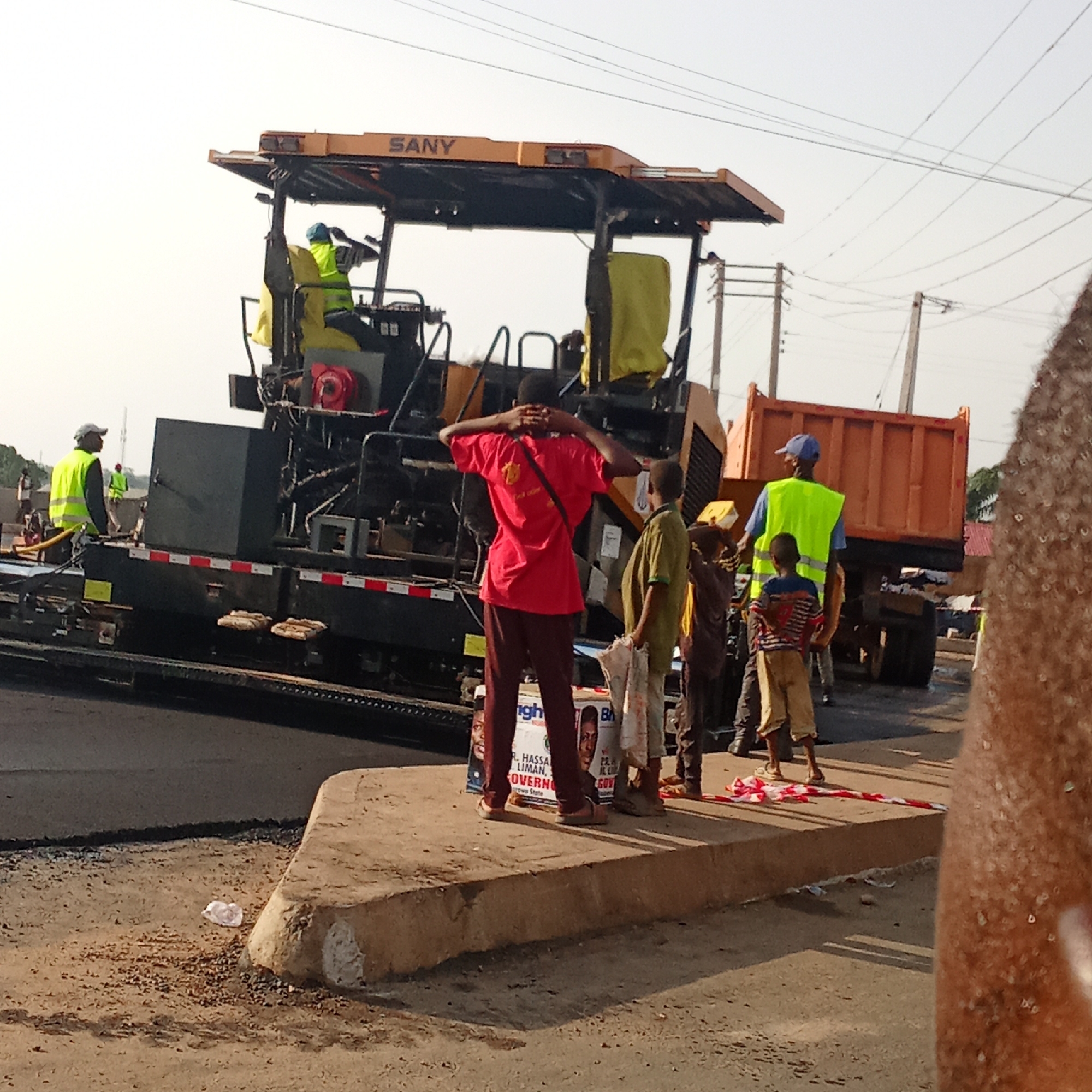 The image depicts road construction taking place in Akwanga, Nigeria. A yellow SANY brand asphalt paving machine is laying new asphalt on the road, operated by a construction worker wearing a yellow vest and blue hat. Another worker in a yellow vest is standing nearby, monitoring the process. A dump truck filled with asphalt is positioned behind the paver. Several people, including children, are standing on the sidewalk, observing the construction. One man in a red shirt is facing the construction equipment, with his hands behind his head. There is a political campaign poster visible on the ground, featuring a photograph of a candidate named “Hugh R. Hassan Liman” with the office “Governo…” and the text “nasawa state”. Red and white striped tape is on the edge of the sidewalk. Utility poles and power lines are visible in the background.