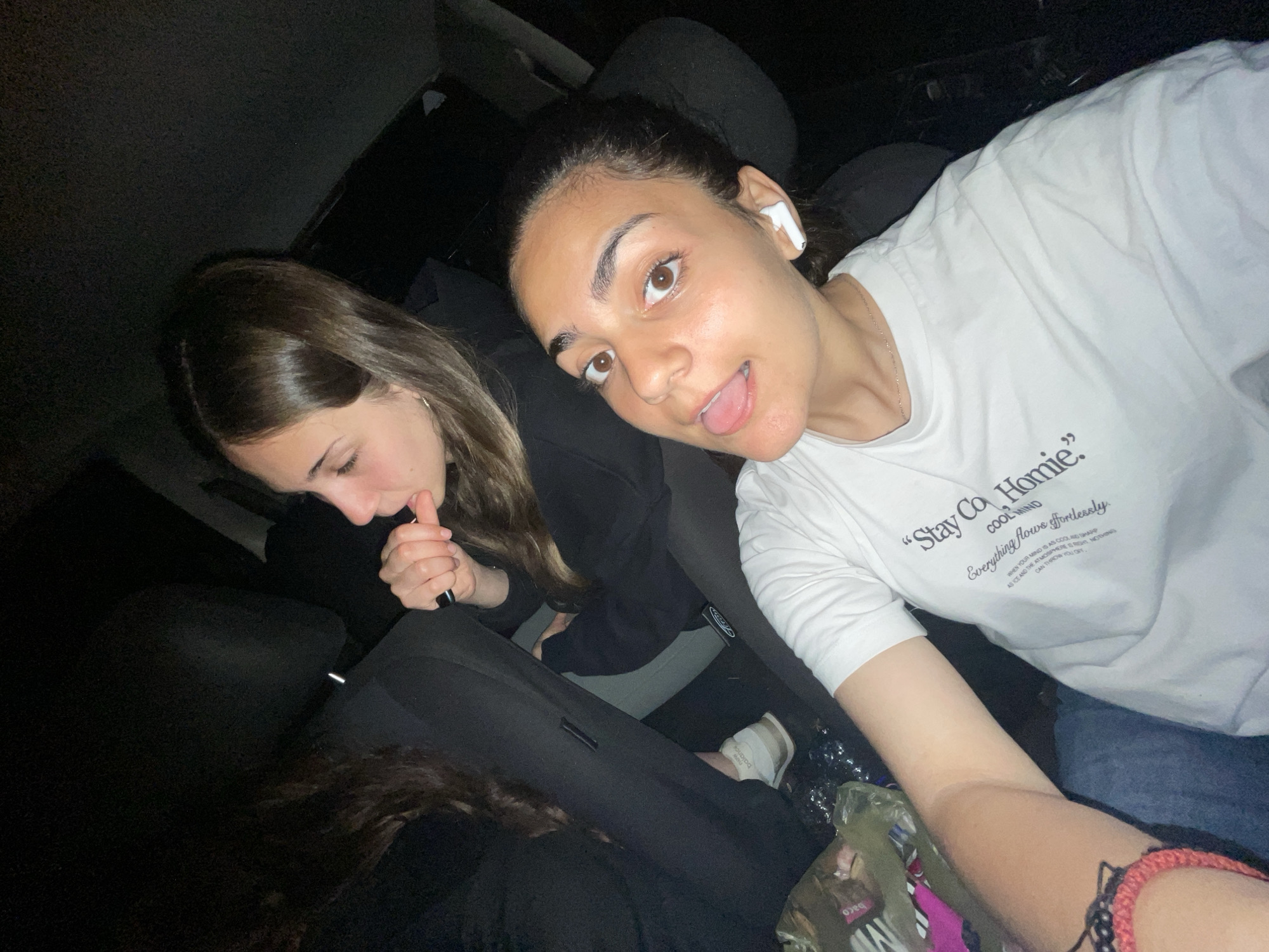 Two young women are inside a car, possibly at night, with limited light. The woman on the right is closer to the camera and is sticking her tongue out while wearing a white long-sleeved shirt with the phrase "Stay Cool Homie". She also has what appears to be an earbud in her right ear. The woman on the left appears to be holding something up to her mouth. The specific location in Debelt, Bulgaria, cannot be determined from the image.