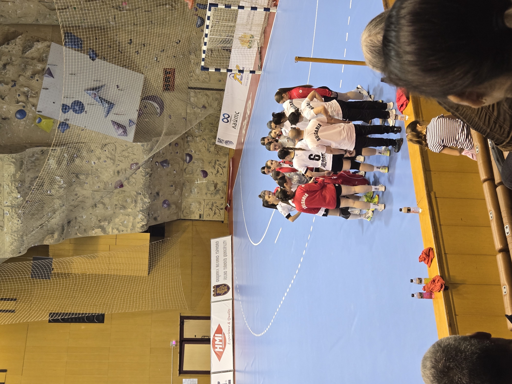 Here is a description of the image:
The image shows a women's handball team, presumably from Bulgaria, gathered in a huddle on a blue handball court. The team is wearing matching jerseys with "BULGARIA" printed on the back. A few players are wearing red tops. In the background, there's a climbing wall with climbing holds, suggesting the handball court might be in a multipurpose sports facility. Also visible are a handball goal, net, and various sports equipment like water bottles and towels. Spectators are partially visible on the sidelines. There is text on the ground showing the name VELIKO TURNOVO LJUNICIPALITY, this may be a location of Veliko Tarnovo, Bulgaria.