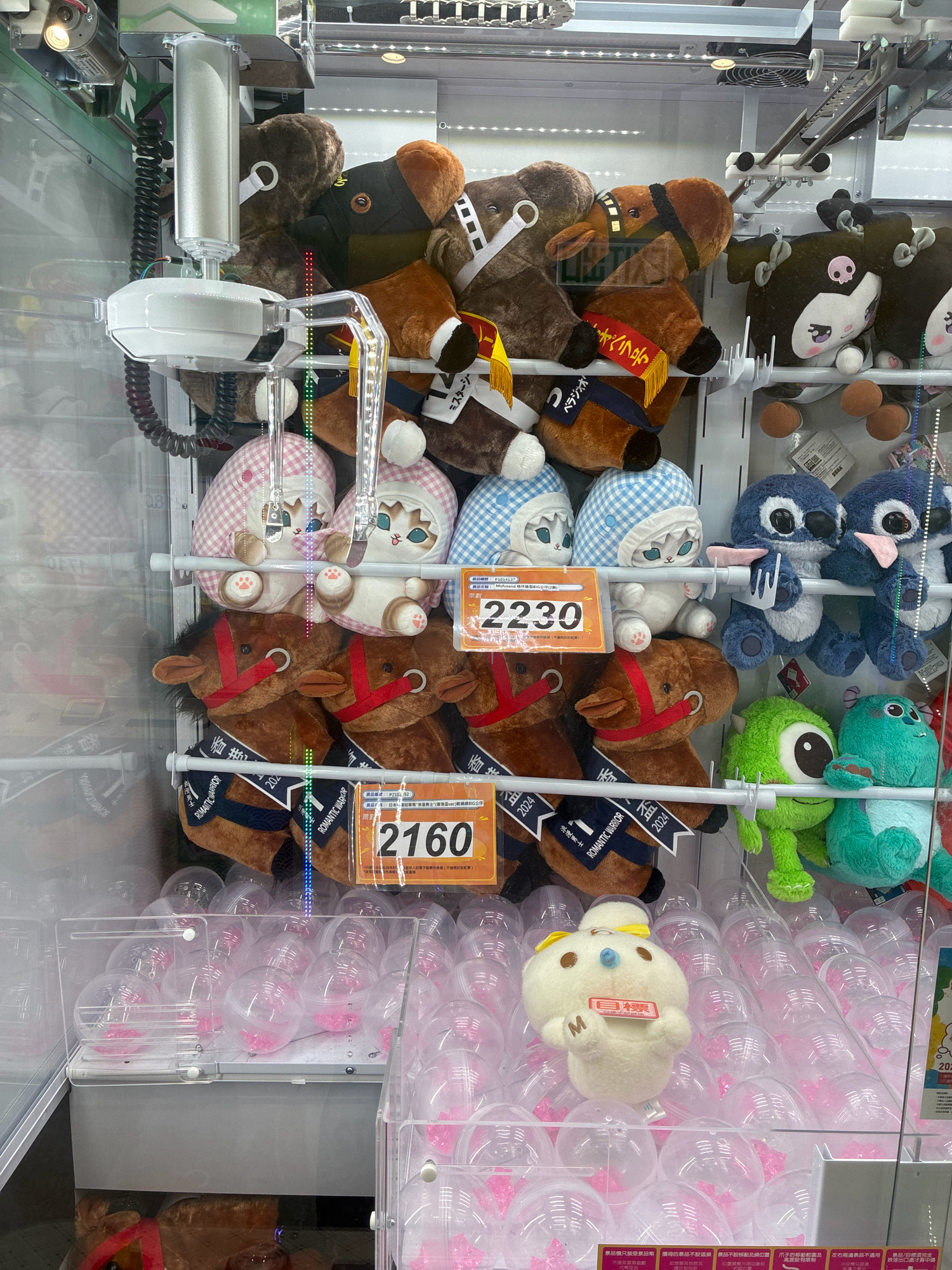 The image shows the inside of a claw machine game located in Hong Kong, China. The machine is filled with various plush toys, including horses, cats, Stitch characters, and what appear to be monsters from the movie Monsters, Inc. In the foreground, there are many clear plastic capsules, some containing pink trinkets. In the center of the machine, a white plush toy with a yellow bow is visible, sitting on top of the capsules. Several price tags are visible on the machine, with the numbers "2230" and "2160" prominently displayed. The overall environment is that of an arcade or entertainment establishment. There are no people visible in the image.