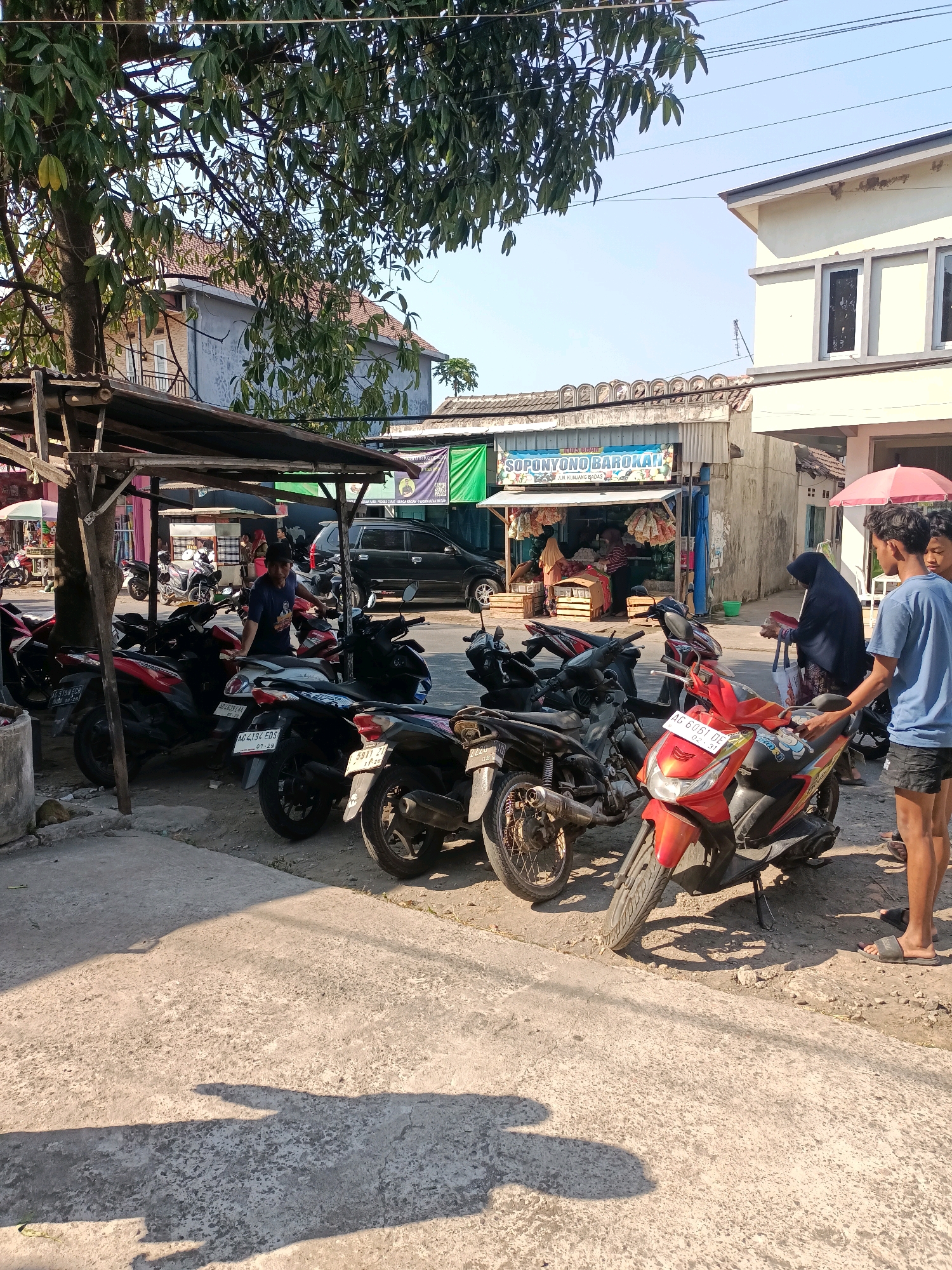 In the city of Badas, Indonesia, a street scene unfolds under a clear, sunny sky, with dappled sunlight casting shadows on a concrete walkway. A row of parked motorcycles, predominantly scooters, lines the dusty roadside, their vibrant colors contrasting with the muted tones of the surrounding buildings and structures. A weathered wooden stall, partially covered by a dark tarp, stands prominently, offering shade to a few of the vehicles.
Behind the motorcycles, a small storefront displays an array of goods, with colorful banners proclaiming "SOPONYONO BAROKAH" and other Indonesian text. Inside, figures are partially visible, suggesting commercial activity. A black SUV is parked further down the street, indicating vehicular presence beyond the motorcycles.
In the foreground, two young men, one in a blue t-shirt and black shorts, the other in a blue t-shirt, stand near a bright orange scooter. To their left, a woman wearing a dark hijab and patterned fabric is seen interacting with another individual partially obscured by her. A pink umbrella is held aloft by someone off-frame, suggesting a solution to the bright sun. The overall atmosphere is one of everyday life in a bustling Indonesian town, with commerce, transportation, and people coexisting on the street.