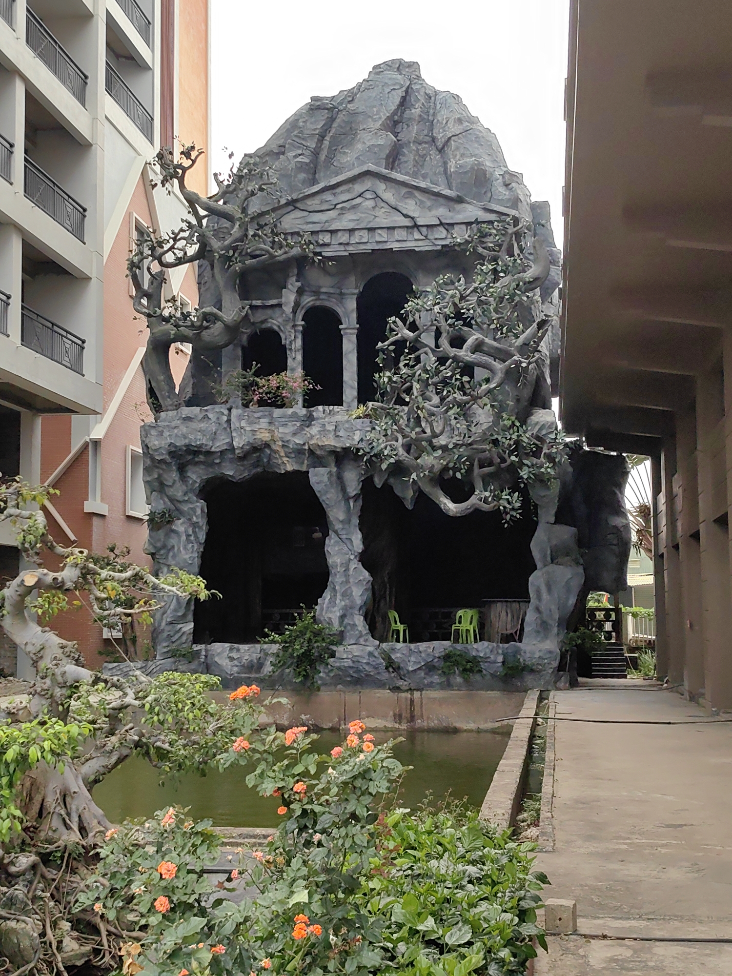 This image shows a rock formation with a building facade located in Tuan Chau Ward, Vietnam. The structure is positioned between two buildings. The formation features a facade with arches and a pediment, designed to resemble a classical architectural style. A pond is in the foreground, surrounded by a variety of plants and flowers, including roses. A narrow concrete walkway runs alongside the pond, leading towards the rock formation and a flight of stairs. Two green plastic chairs are inside the rock formation. The buildings on either side of the structure are modern, multi-story buildings.