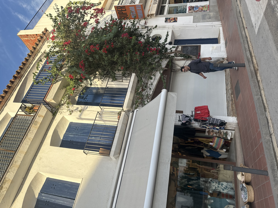 This image depicts a daytime street scene in Calella De Palafrugell, Spain. The foreground features a brick-colored paver sidewalk and a gray asphalt street.
On the left, a two-story building with white stucco walls is visible. The ground floor houses a retail establishment with a large glass display window, showcasing mannequins dressed in swimwear and clothing. Baskets containing merchandise are placed outside, and racks display red shorts and various swimwear. A light-colored retractable awning extends over the storefront. Above, two windows with closed dark blue wooden shutters and dark metal railings on small balconies are visible.
To the right of this, a large, vibrant bougainvillea plant with numerous red-pink flowers and green foliage climbs the white wall of the building, partially obscuring a third window with dark blue shutters and a balcony. Beneath the bougainvillea, a white roller shutter indicates a closed establishment, with the word "restaurant" faintly visible above.
Further right, another storefront is partially visible, featuring a sign with orange text that reads "SUPER MERCADO PANADERIA" (Supermarket Bakery). The upper levels of the buildings feature additional balconies with dark railings and visible terracotta roof tiles. The sky is clear blue with sparse white clouds.
A single male subject is present, walking on the sidewalk from right to left in the mid-ground. He is a middle-aged man with dark hair and glasses, wearing a dark gray long-sleeved shirt or hoodie and blue jeans. He is looking forward and slightly to his left. A dark gray bollard is positioned on the sidewalk to his left. Bright sunlight illuminates the scene, casting shadows.