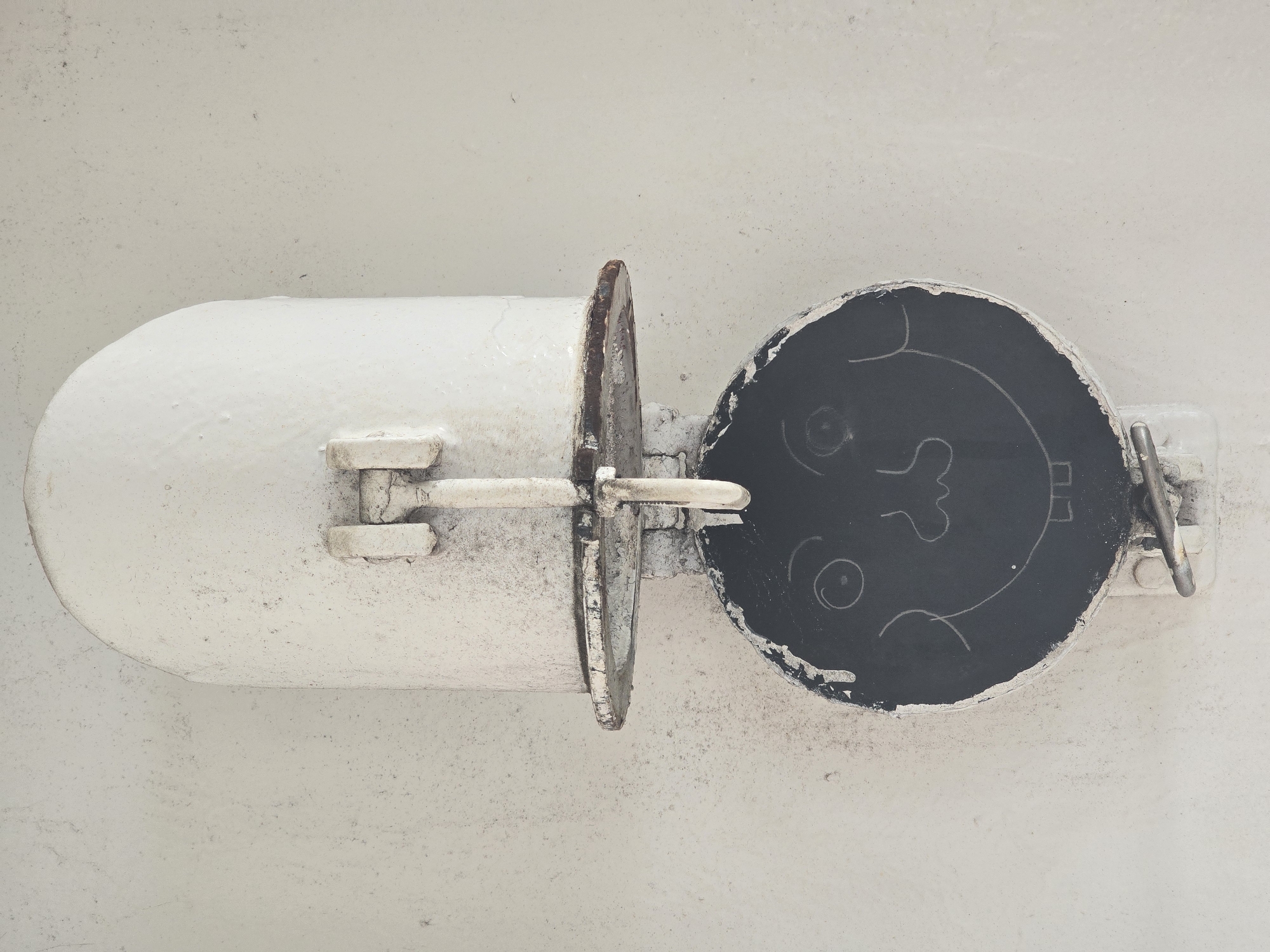 The image shows a close-up, head-on view of a white, weathered metal fixture attached to a textured, light-colored wall. The fixture has a rounded top and a circular opening below it. A crude drawing of a face with wide eyes, a nose, and a smiling mouth with two teeth is visible within the circular opening. The drawing appears to be made with chalk or a similar marking material. There are metal brackets and hinges on the fixture, suggesting it may be a functional part of a larger structure, possibly ventilation or an access point. The overall impression is one of an industrial or utilitarian object that has been playfully altered with the addition of the drawing. The location is specified as Unknown, United Kingdom.