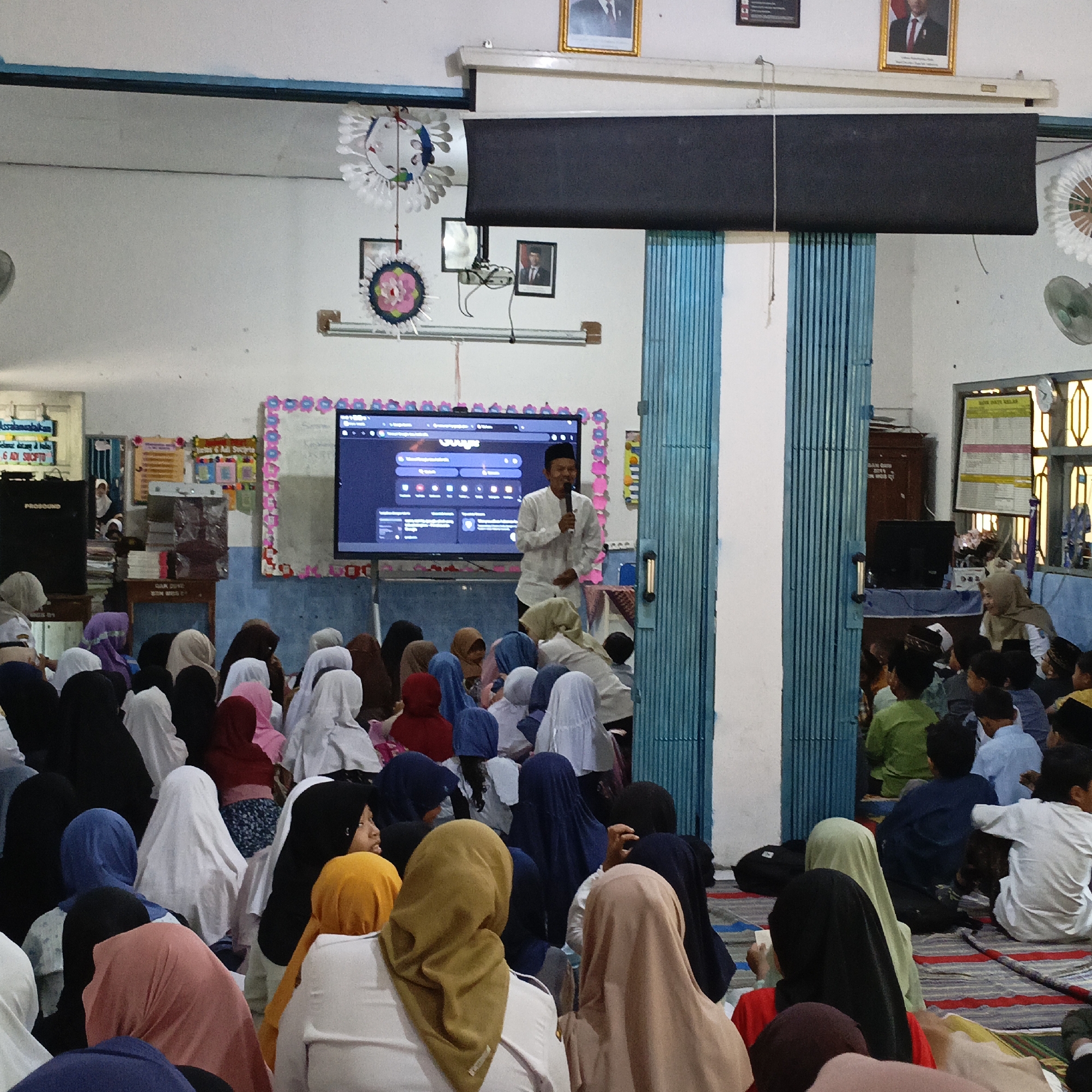 The image depicts an indoor gathering of approximately 40-50 people, mostly children and young adults, seated on the floor in a room. A man in white attire is standing and speaking into a microphone in front of a large screen. The screen displays a presentation, likely educational or religious. The room appears to be a classroom or community hall, with decorations on the walls, including framed pictures and colorful banners. The setting is consistent with a school or religious institution in Salatiga, Indonesia, given the cultural attire and the presence of a projector screen. The atmosphere seems to be one of learning or communal activity.