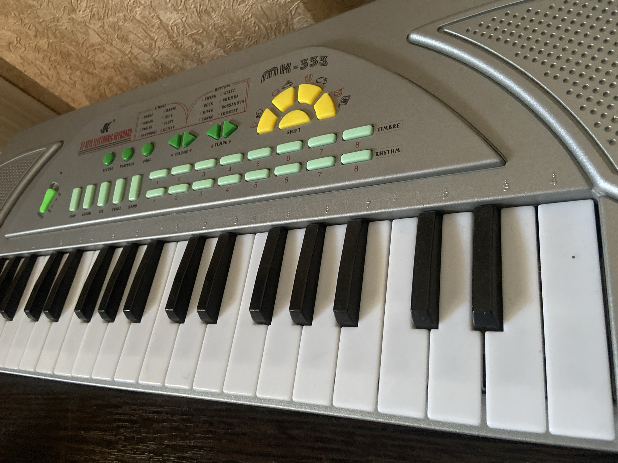 The image shows a partially visible, silver-colored MK-333 37-key electronic keyboard. The keyboard has black and white keys, with various control buttons above them, including green buttons labeled "RECORD," "PLAYBACK," "PROG," "STOP," "CHORD," "GUIDE," and "DEMO." There are also yellow "SHIFT" buttons, and green buttons for volume, tempo, timbre, and rhythm. The background appears to be a wooden surface. There is no evidence to suggest that this image was taken in the city of Kazan, Russia.