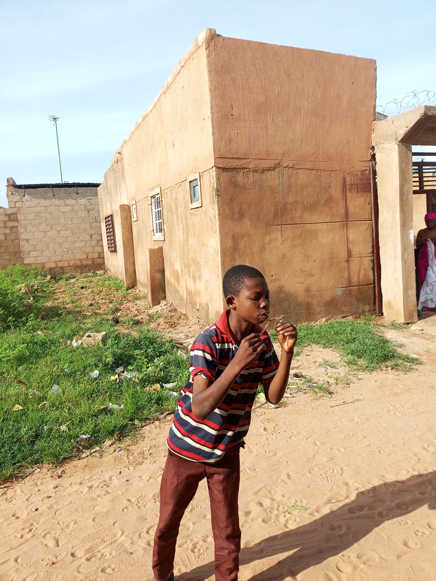 A young boy wearing a striped shirt and brown pants stands in a sandy area in front of a mud-brick building. He is holding a small twig to his mouth. To the right of the boy, a woman in a pink headscarf and white garment stands near a doorway, holding a piece of fabric. The building appears to be in disrepair, with some sections of brickwork exposed. The ground is a mix of sand and patchy green grass, with some scattered debris. The sky is a clear blue. The location is identified as Tempure, Nigeria.
