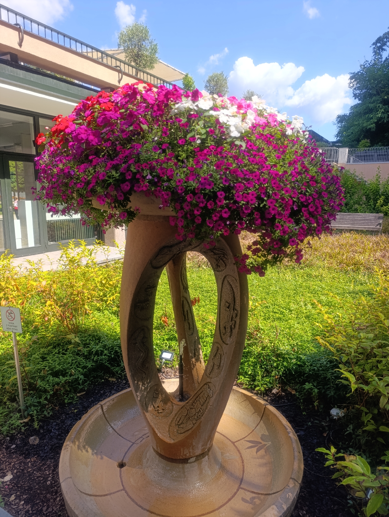 This is a medium shot photograph of a decorative fountain with abundant flowers, located in Luhačovice, Czechia. The fountain itself is made of a light brown, textured material and features an oval opening with carved embellishments. It is filled with water in its basin. Overflowing from the top of the fountain is a lush arrangement of colorful flowers, predominantly in shades of pink and magenta, with accents of white and red. The fountain is set in a garden environment with green grass and various types of vegetation. In the background, there is a building with a covered walkway, a railing, trees, and a park bench. A sign with text is visible to the left of the fountain. The sky above is blue with scattered white clouds.