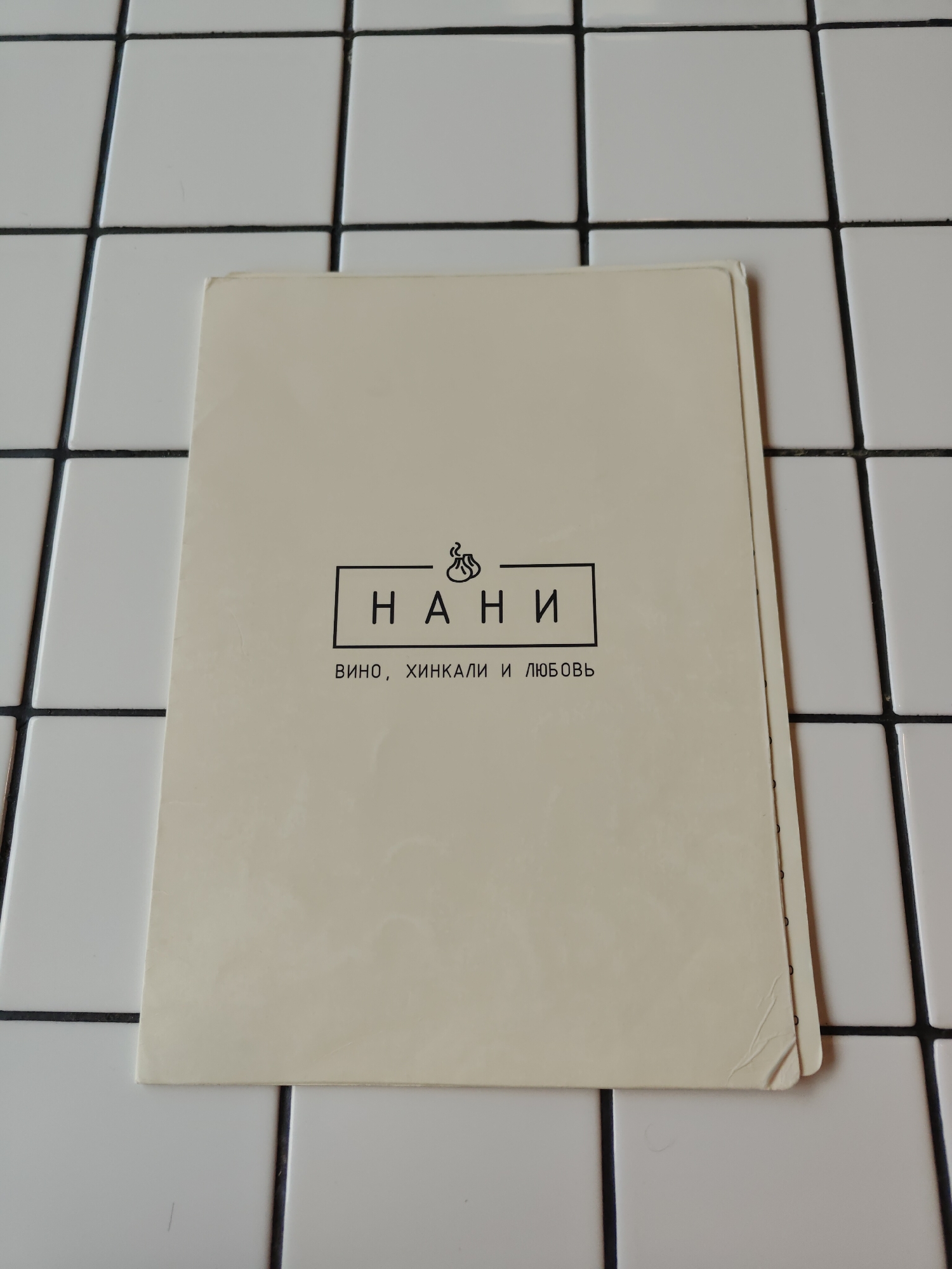 A close-up, top-down shot features a cream-colored menu card placed on a white tiled surface with black grout lines. The menu card is positioned diagonally across the frame, covering the lower half of the image. The top portion of the card displays a stylized logo that appears to be a steaming dumpling or khinkali. Below the logo, the word "НАНИ" is prominently displayed within a black rectangular border, written in sans-serif capital letters. Beneath this, the text "ВИНО, ХИНКАЛИ И ЛЮБОВЬ" is printed in a smaller, sans-serif font. The tiled surface is clean and well-lit, with the black grout creating a grid pattern. The menu card has a slight crease on its lower right corner. The scene is devoid of any people or explicit indication of location in Кварса, Russia, beyond the general setting of a menu on a tiled surface.