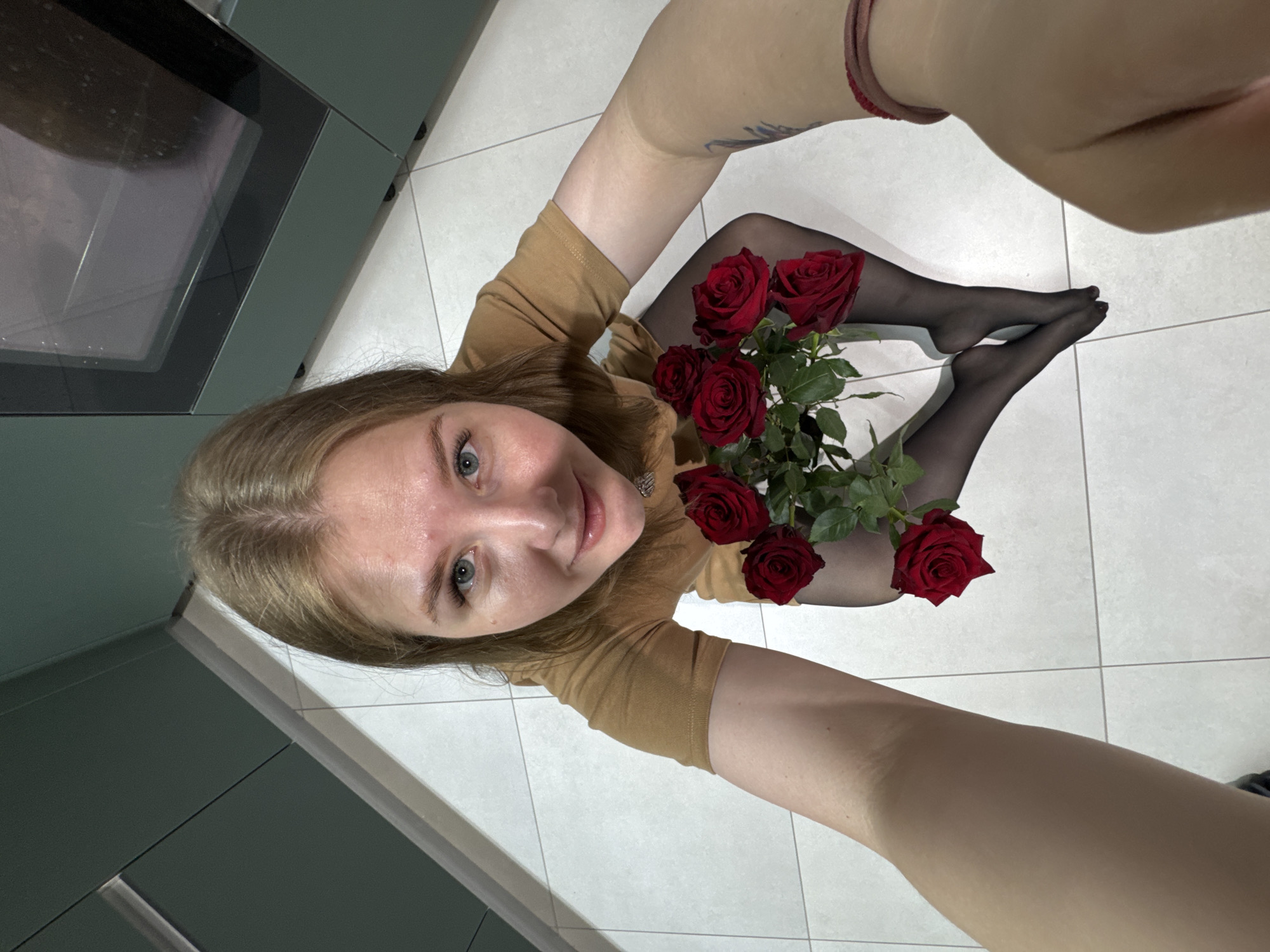 A woman is taking a selfie in a kitchen. She is wearing a tan long-sleeved shirt and black tights. She is holding a bouquet of red roses between her legs and feet, which are also clad in black tights. The kitchen has white tile flooring and modern green cabinets. The scene is likely indoors. The location is Gornyi (Dvurechenskoe S/P), Russia.