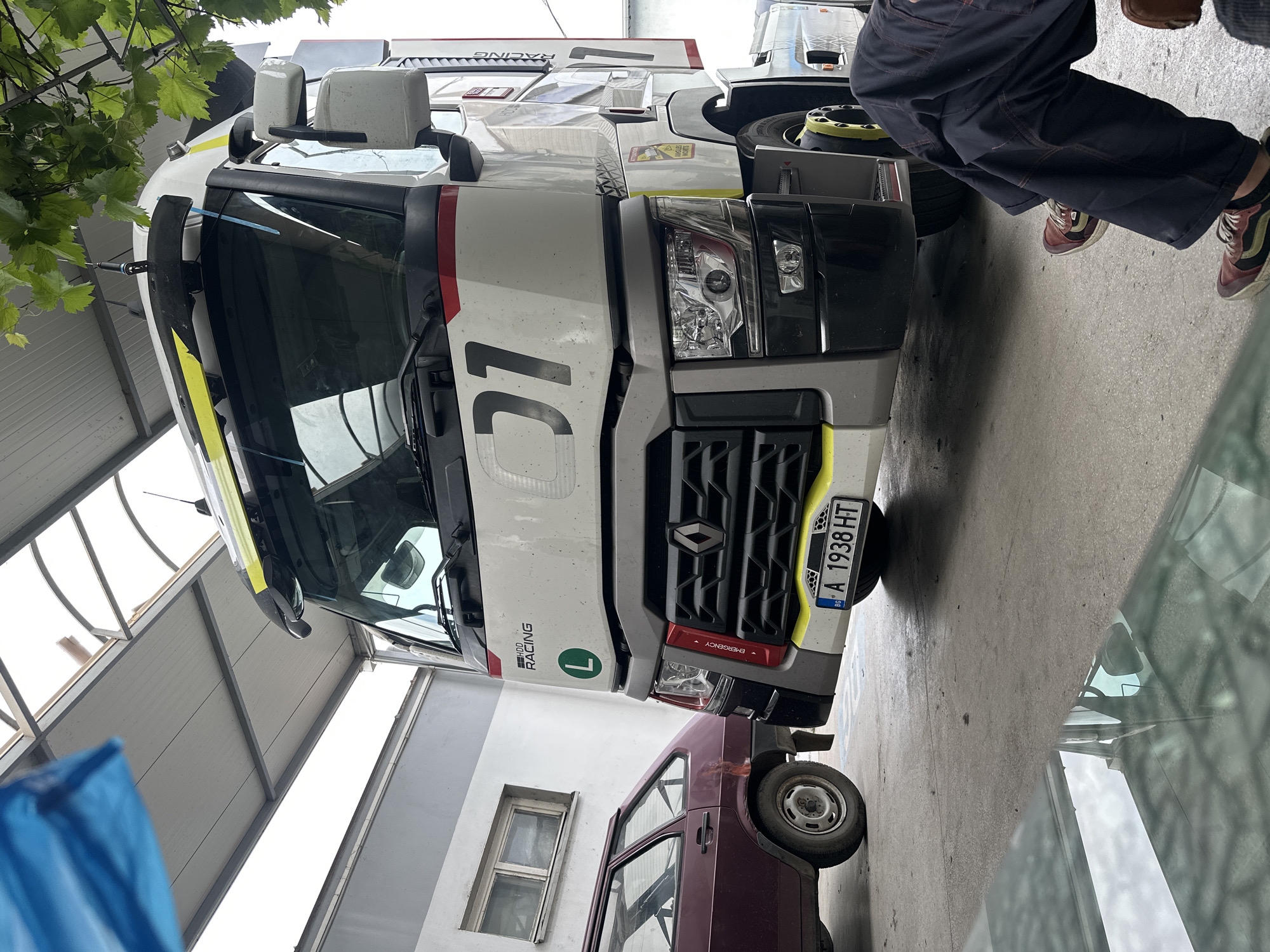 A white semi-truck, likely a Renault model given its grille logo, dominates the central view. It features racing livery, including a large "01" on its front, "RACING" text, and "HDD RACING" on its side. A green 'L' symbol is visible near the grille, and a small "EMERGENCY" label is present near the headlights. The truck's license plate, "BG A 1938 HT," indicates its origin.
A smaller, maroon-colored car is partially visible, parked in front of the truck. To the right, the lower half of a person is seen, wearing dark pants and multi-colored sneakers. The scene takes place in what appears to be a garage or workshop, characterized by a concrete floor, white walls, and bright overhead lighting from skylights. Green vine foliage hangs into the upper left corner of the frame. A blue object or bag is partially visible in the bottom left foreground. The time of day appears to be bright daytime. The overall scene is static, showing vehicles parked in an indoor setting.