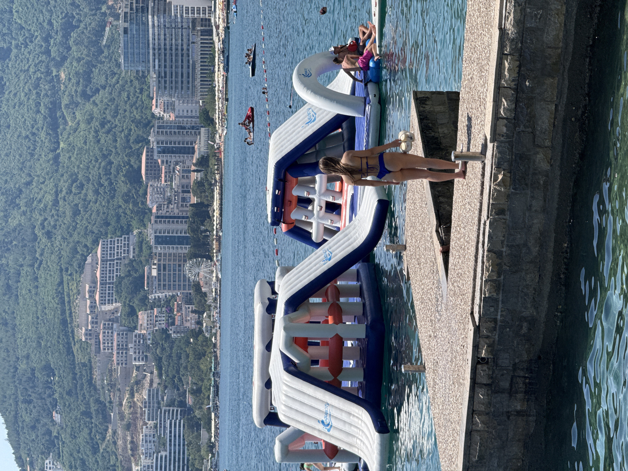 In Budva, Montenegro, on a bright, sunny day, a woman in a blue bikini stands on a stone pier, holding a white hat, looking out at a floating inflatable water park in the sea. The water park features large, white and blue inflatable structures with slides and climbing elements. Several people are visible on and around the water park, some swimming, others relaxing on inflatables. In the background, the cityscape of Budva rises up a green, forested hill, with numerous buildings, hotels, and a Ferris wheel visible. The sea is calm and reflects the blue sky.