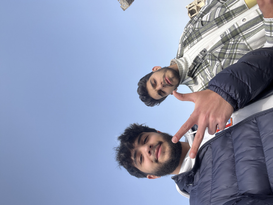 Joubar Municipality, Syria: A selfie-style image depicts two young adult males outdoors under a clear, light blue sky. The male in the foreground, positioned to the left, has dark curly hair and a full beard, wearing a dark blue puffer jacket over a white hooded sweatshirt. He is making a 'V' gesture with the index and middle fingers of his right hand and has a slight smile. The second male, to the right and slightly behind, has dark hair and a short beard, wearing a white ribbed turtleneck under a green and white plaid shirt. He holds a light-colored cup in his right hand, partially visible at the bottom right, and has a neutral expression. In the far background on the right, partially destroyed or derelict building structures are visible against the sky, indicating a damaged urban environment.