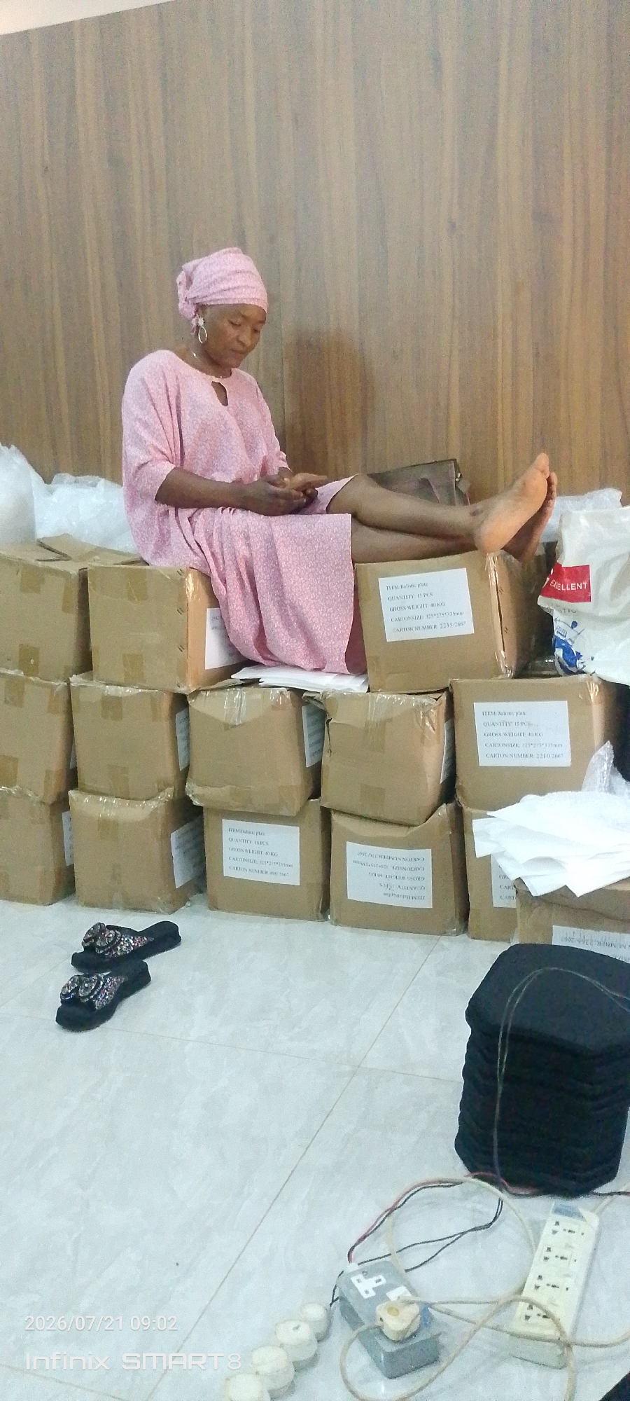 A woman, appearing to be of African descent, is sitting on a stack of cardboard boxes in what appears to be a storage area. She is wearing a light pink headscarf and a matching pink patterned dress. Her legs are crossed and extended, and she is looking down at a mobile device she is holding in her hands. Her feet are bare. The boxes are stacked neatly in multiple rows, with some boxes having labels that are partially visible. In the foreground, there is a pair of black and sparkly flip-flops. To the right of the boxes, there is a black, cylindrical object, possibly a fabric roll, with wires and electrical components, including a power strip and what looks like a small battery pack, scattered on the floor. The background consists of a wooden-paneled wall. The floor is tiled. A timestamp in the lower left corner indicates the photo was taken on July 21, 2026, at 09:02. Based on visual cues like the woman's appearance and the potential context suggested by "smart" technology, and the likely region where this type of storage and everyday scene might occur, this scene is consistent with a location in Kakau, Nigeria.