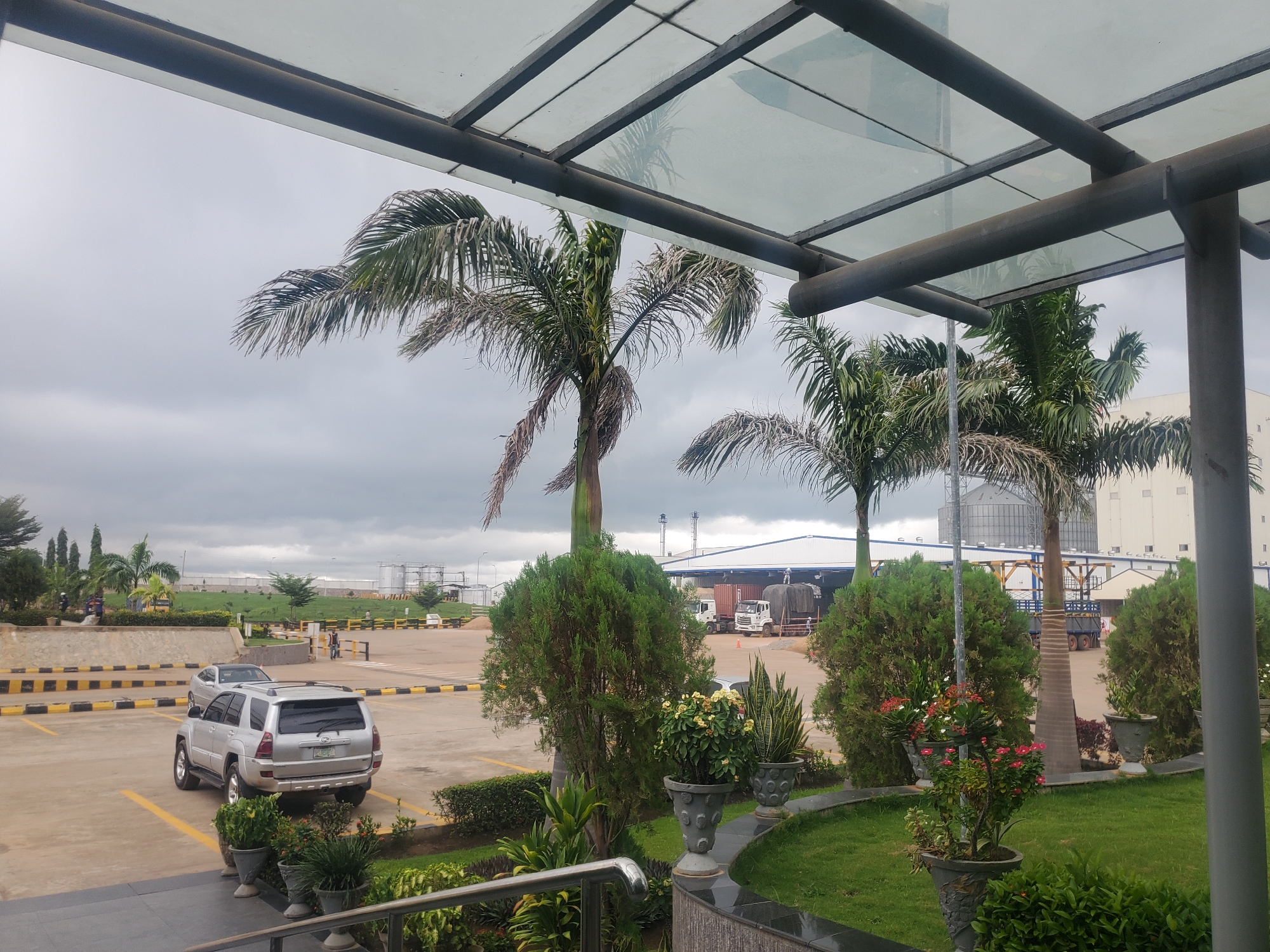 The image depicts an outdoor scene in Gwagwada, Nigeria, under an overcast sky. In the foreground, a silver SUV is parked in a lot marked with yellow and black lines. Beyond the SUV, palm trees and manicured bushes are visible. In the middle ground, there is a large industrial building with a white facade, featuring large silos and several trucks parked in front of it. A person can be seen walking in the distance. The overall environment suggests an industrial or commercial area with landscaping.