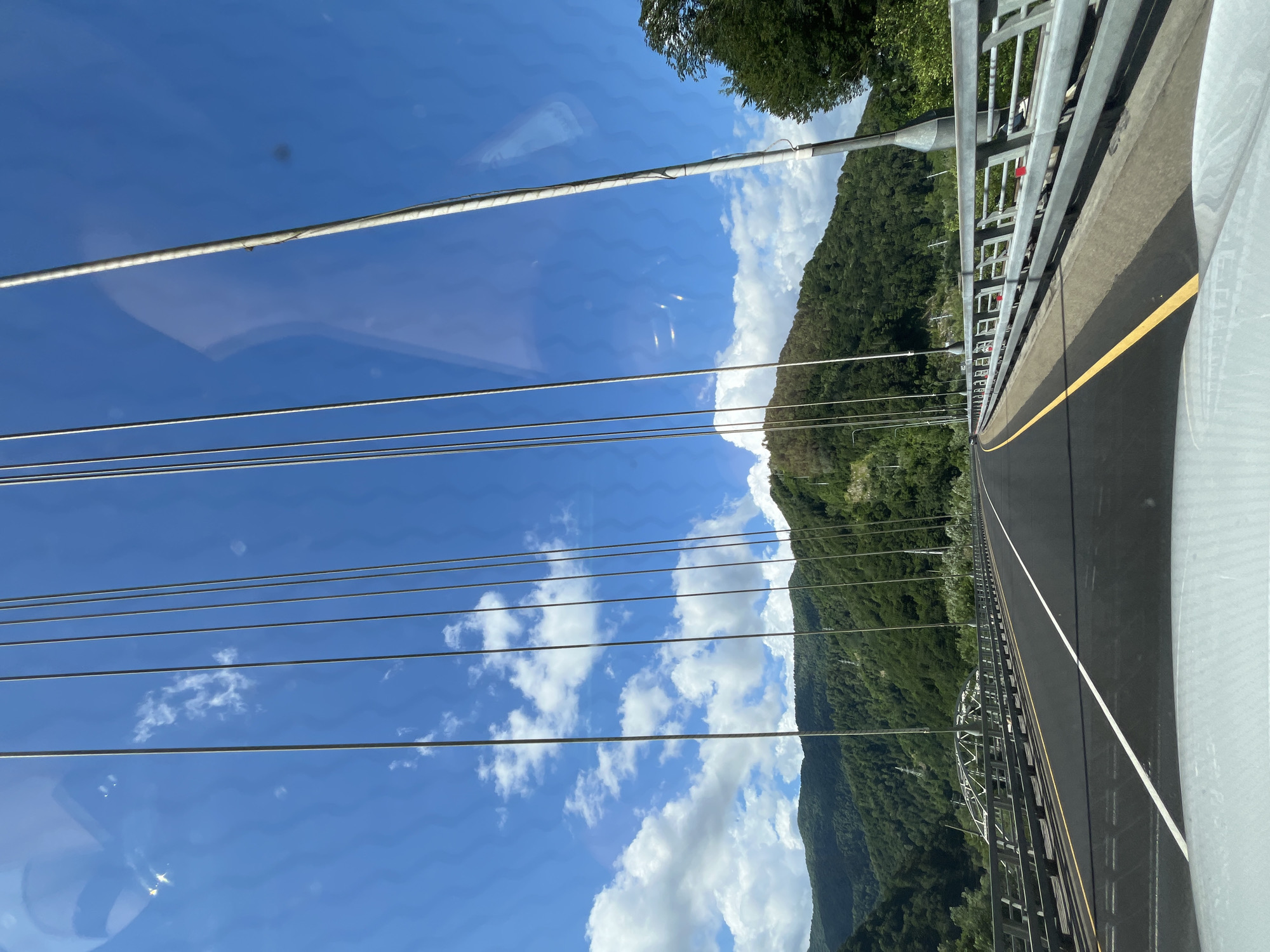 The image captures a view from a vehicle driving across a modern suspension bridge on a clear, sunny day. The bridge has a paved roadway with white and yellow lane markings. Along the sides of the road are metallic guardrails. Numerous vertical cables, characteristic of a suspension bridge, extend upwards into the bright blue sky, which is dotted with fluffy white clouds.
In the background, lush green, tree-covered mountains rise steeply. Some of these mountains show signs of human activity, such as what appear to be power lines or ski lifts traversing their slopes. The bridge itself is set within a mountainous valley.
The location is identified as the bridge over the Psezuapse River in Kepsha, Russia, which is part of the Sochi area. The environment appears to be a natural landscape with significant infrastructure. There are no visible people or animals in the frame, only the man-made structures of the bridge and the natural beauty of the surrounding mountains and sky. The reflections on the car's windshield are also a prominent feature of the image.