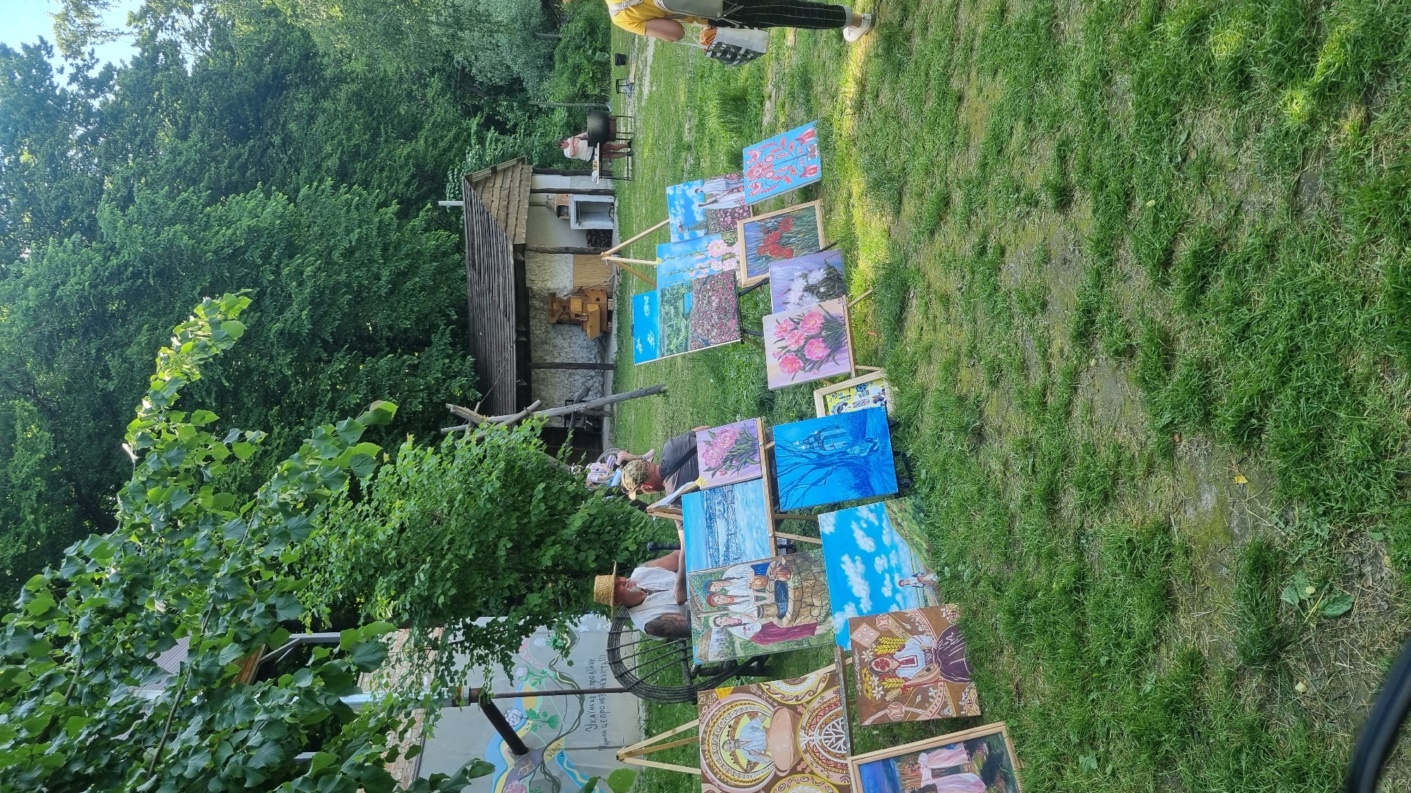 This image captures an outdoor art display in Melnyky, Ukraine, featuring multiple painters and their artwork set against a backdrop of traditional buildings and dense foliage.
In the mid-ground, a man with a light-colored straw hat and an embroidered white shirt is seated on the left, conversing with another man in a dark cap and t-shirt. Both men are positioned near a collection of paintings displayed on wooden easels and directly on the grassy ground. The paintings exhibit diverse subjects including stylized portraits, traditional Ukrainian scenes (women at a well, figures in national attire), landscapes (snowy village, sunny fields), and floral arrangements (peonies, poppies). A wall with a painted mural featuring text in Ukrainian and Russian is visible behind the seated men.
To the right, a small traditional wooden building with a brown shingled roof and white-washed walls is present. A woman in a light-colored top stands next to a large dark pot placed on a stand, suggesting food preparation, with a table nearby. Another individual, wearing a yellow shirt and dark striped pants, is partially visible on the far right, walking away from this area. More paintings are displayed on easels in this section of the grassy expanse.
The foreground consists of green grass with some worn patches. The background is dominated by a dense wall of various green trees and foliage, with a sliver of clear sky visible at the upper right. Several unidentifiable wooden structures are scattered throughout the scene. The overall lighting suggests bright daylight.