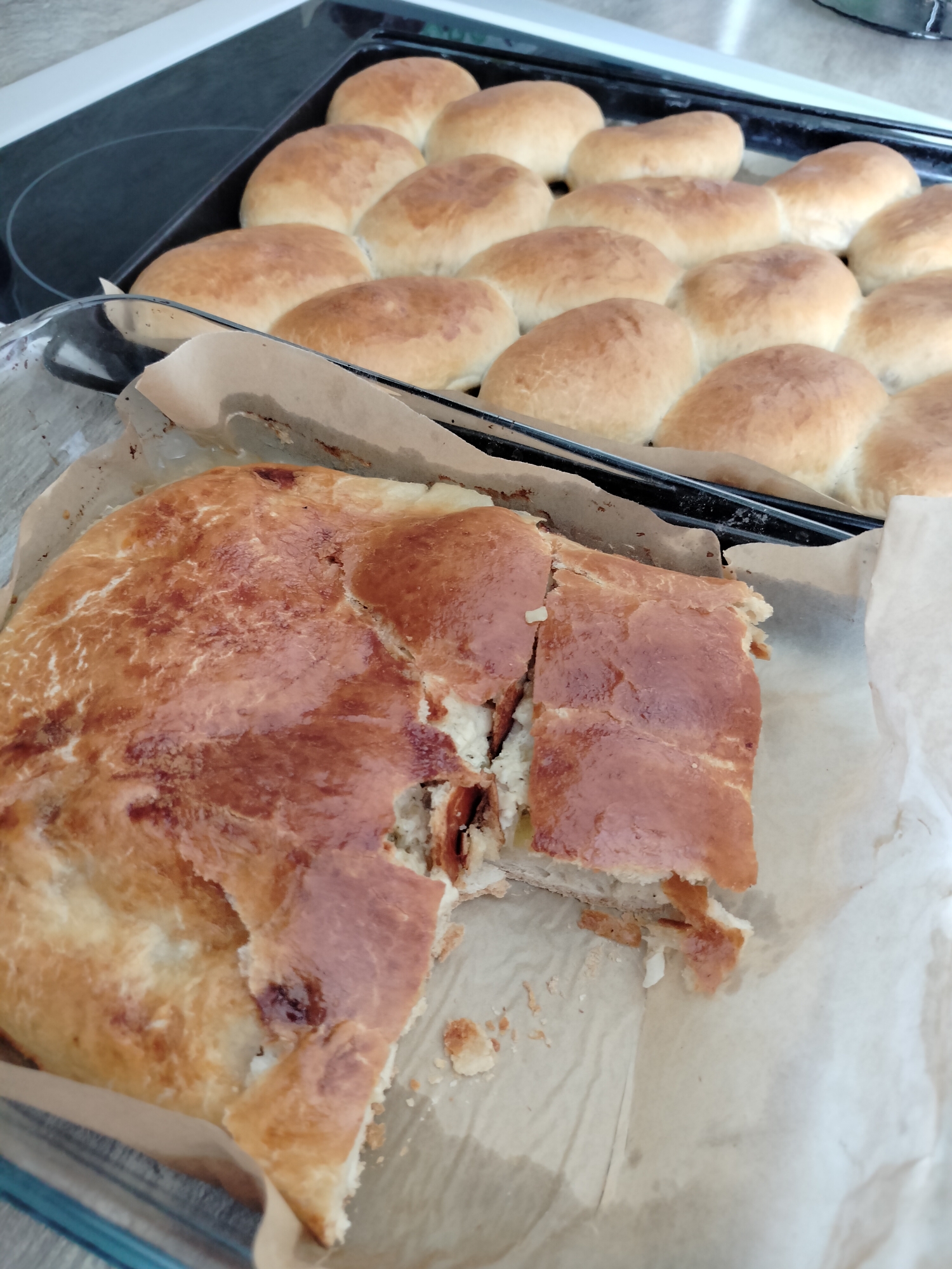 A close-up, top-down view captures a baked pie with a golden-brown crust, partially sliced, revealing a light-colored filling. The pie rests on parchment paper within a clear glass dish. In the background, a baking sheet holds a row of small, golden-brown baked rolls, also on parchment paper. The scene is set on a light-colored countertop, with a dark stovetop visible to the left. This image was captured in Yashkino, Russia.