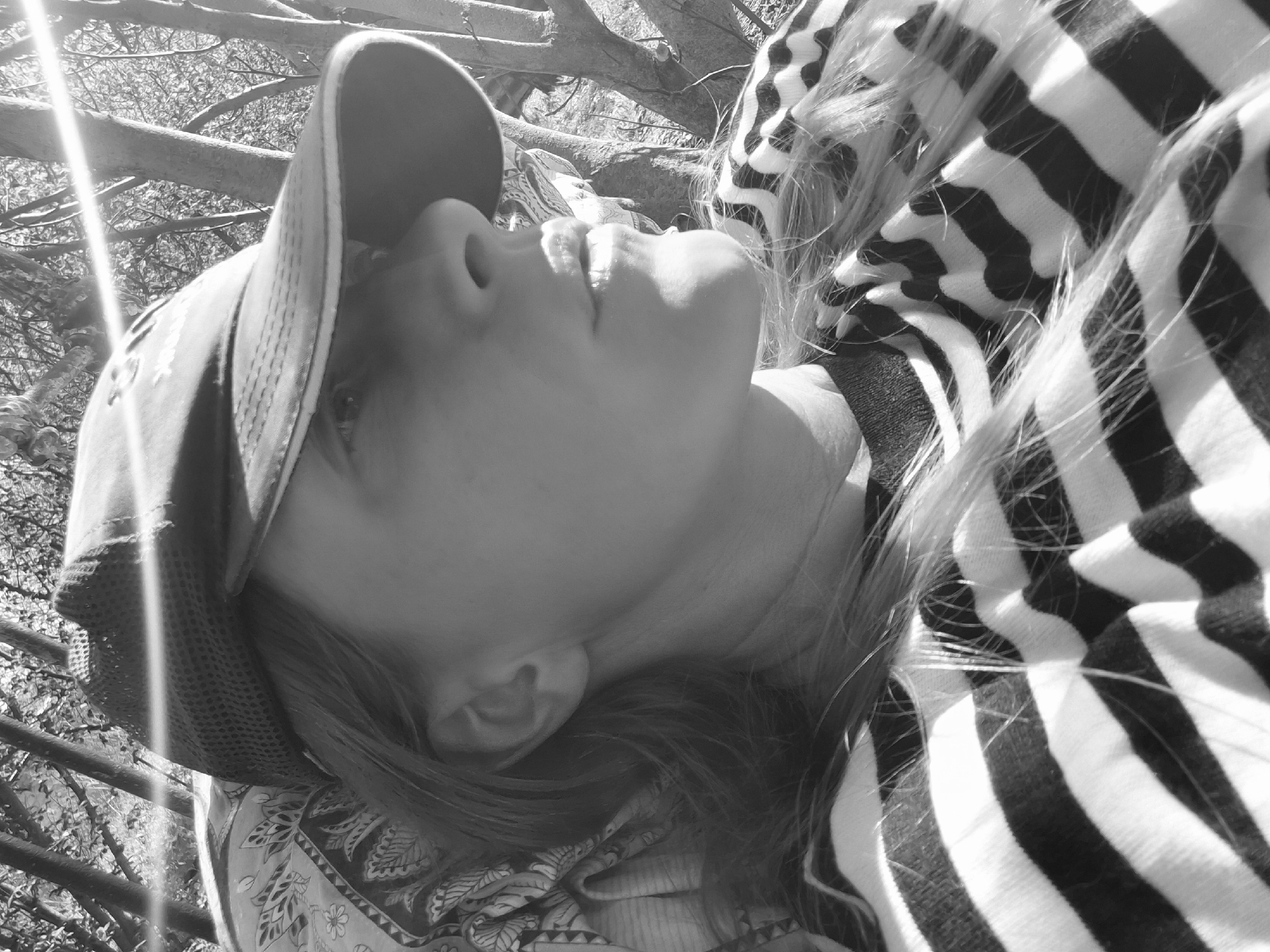 A grayscale image shows a person wearing a baseball cap, striped shirt, and patterned garment. The person lies on their back, looking upwards towards tree branches overhead, with sunlight filtering through. No location is discernible.