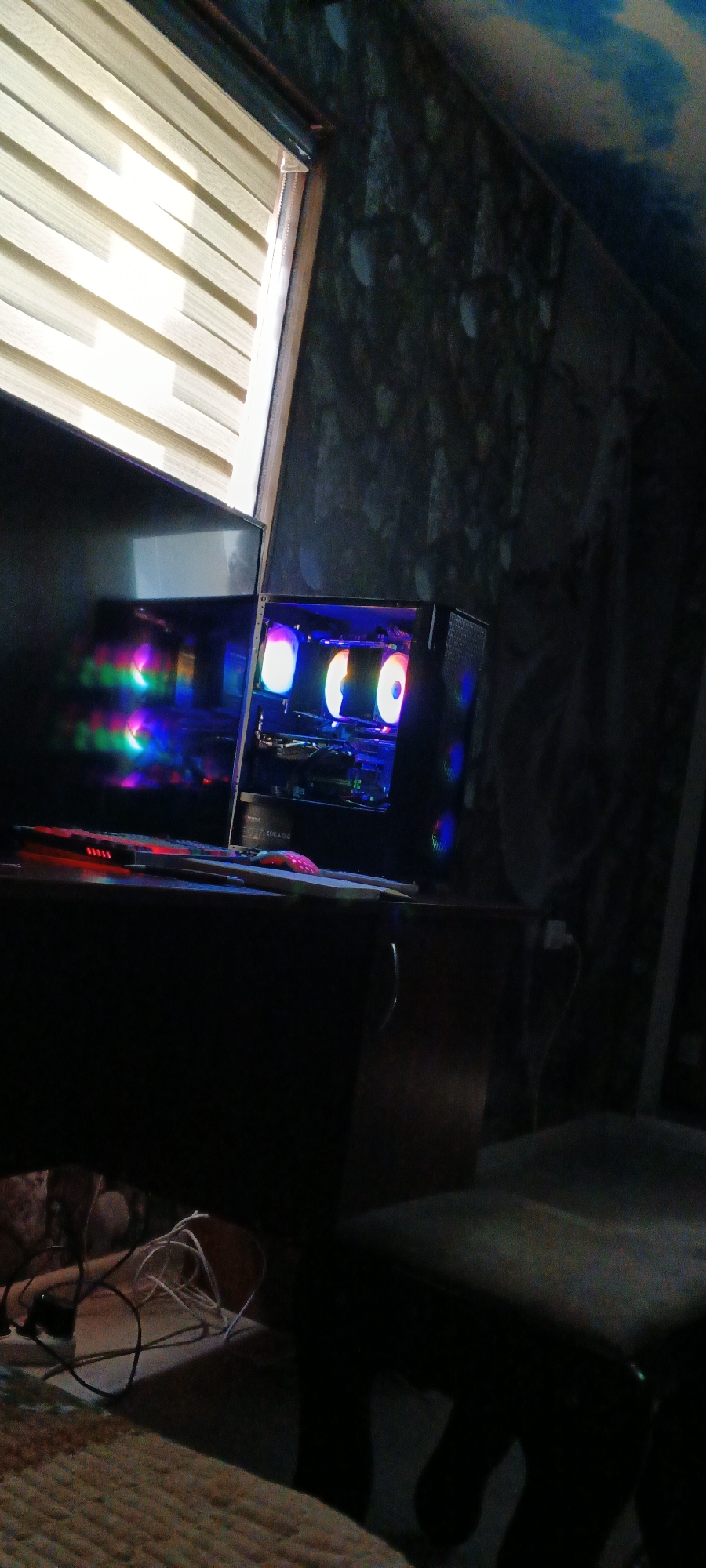 In an indoor setting, possibly a bedroom or office, a computer setup is visible on a desk. A black computer tower with multiple RGB fans emitting colorful lights is on the right side of the desk. To the left of the computer tower, a monitor is positioned, reflecting the RGB lights from the computer. In front of the monitor and computer, a keyboard with red backlighting and a pink mouse are on the desk. The desk appears to be made of dark wood. Behind the desk, a wall with a textured, dark gray pattern is visible, and a portion of the ceiling shows a blue and white cloud-like mural. A window with horizontal blinds that are partially open allows light to enter the room. Various cables are scattered on the desk and on the floor below the desk. The floor in the foreground is covered with a textured rug. The overall lighting in the room is dim, with the primary light sources being the RGB lights from the computer and the light coming through the blinds. There are no people visible in the image. The scene does not provide any information to indicate a location in Mustakillik, Uzbekistan.