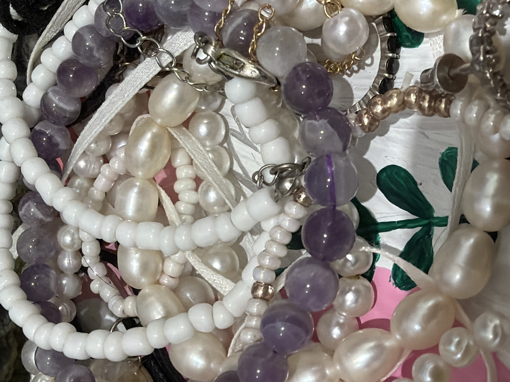 The image shows a close-up view of a collection of jewelry. There are multiple strands of beads, including round purple beads, irregular white pearls, and small white round beads. Some strands are connected with silver clasps and chains. There are also ribbons and bits of other jewelry pieces mixed in. The background features hints of pink and white with some green patterns.
The image does not appear to be connected to Izhevsk, Russia.