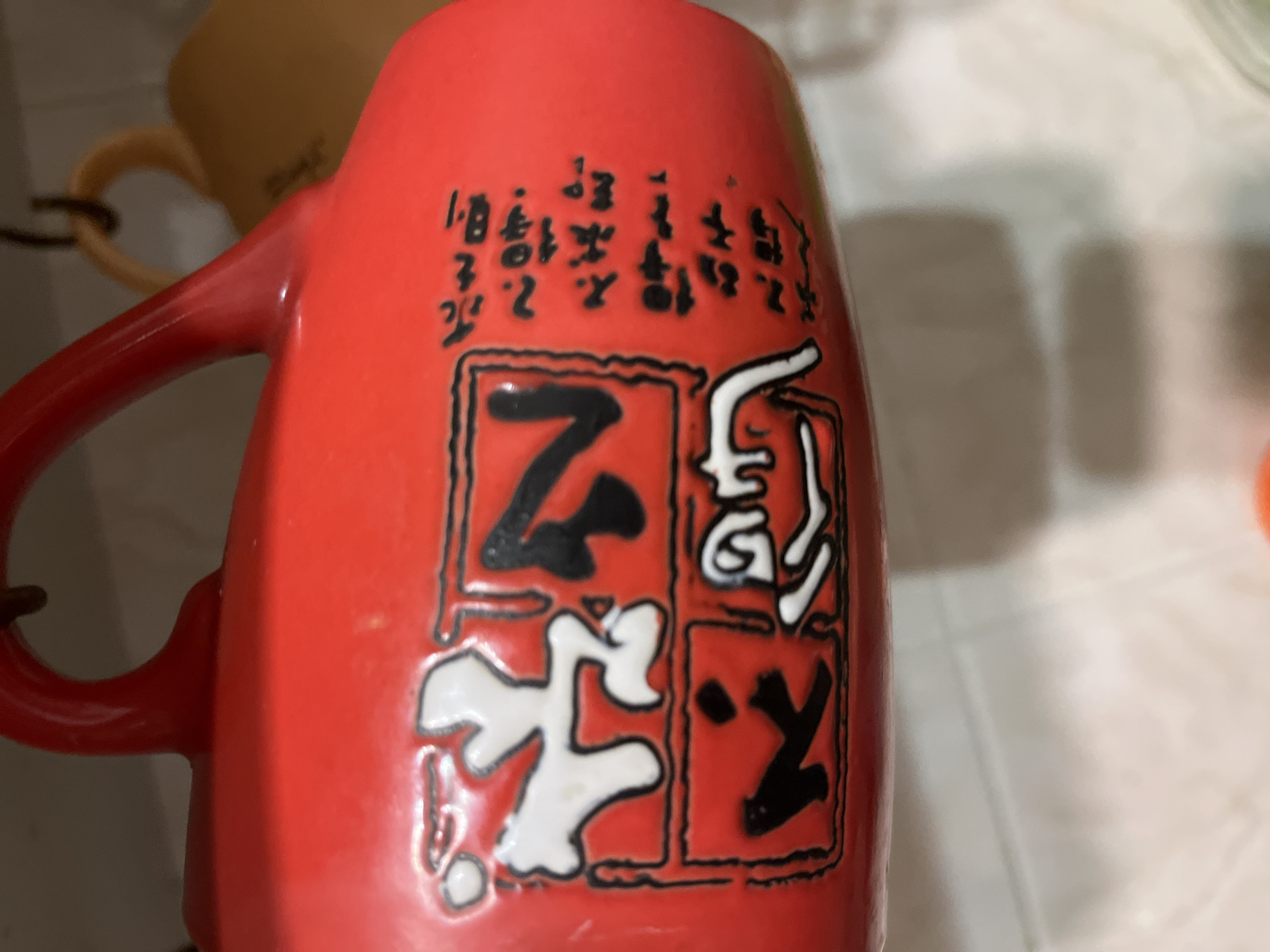 The image shows a close-up of a red ceramic mug with Japanese characters printed on it. The characters are enclosed in black outlines and appear to be in white and black. To the right of the characters, there is vertical text also in black. The mug has a red handle. The background is blurred and appears to be a light-colored tiled surface. The scene is indoors, likely a kitchen or dining area. The mug is hanging from a hook. There is no indication of location in Minsk, Belarus.