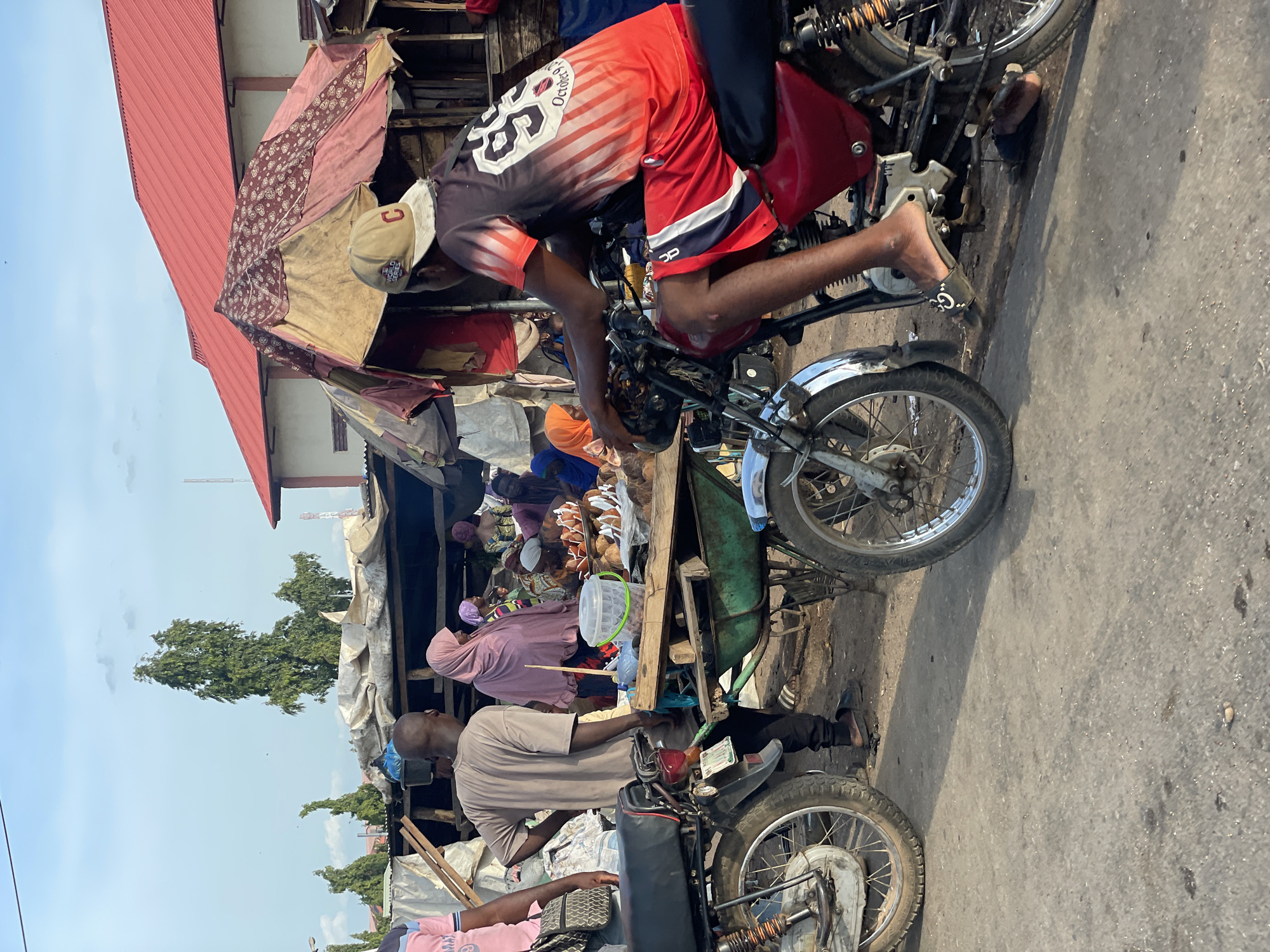 The image depicts a bustling street scene in Lokoja, Nigeria, likely a market area. Several people are present, including vendors and passersby. One man is positioned on a motorcycle, inspecting or adjusting something near the handlebars. Other motorcycles are also visible in the foreground, along with a hand cart near a table. Vendors are behind tables with various goods. Several buildings with red roofs and tarpaulins are in the background, indicating a commercial or residential area. The environment is urban with signs of street activity.