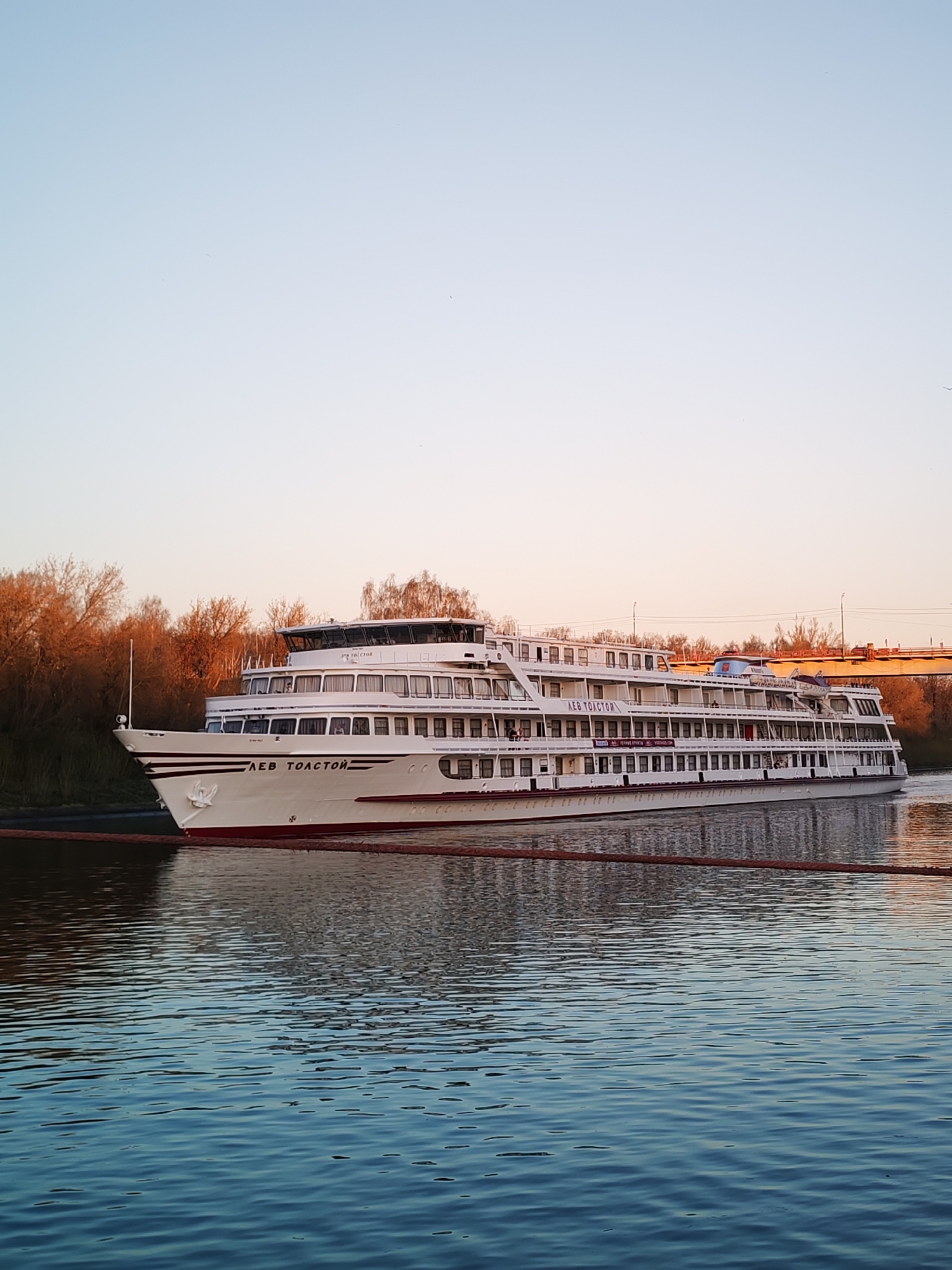 The image depicts the cruise ship "Lev Tolstoy" on the Yakhroma Canal in the city of Yakhroma, Russia. The ship is white with several decks and has the name "LEV TOLSTOY" written on its side in Cyrillic. A brown safety rope is visible in the foreground. The water in the canal is rippled and reflects the ship and the sky. In the background, there are trees with orange foliage and a bridge. The sky is a gradient of light blue and pink, indicating sunrise or sunset.