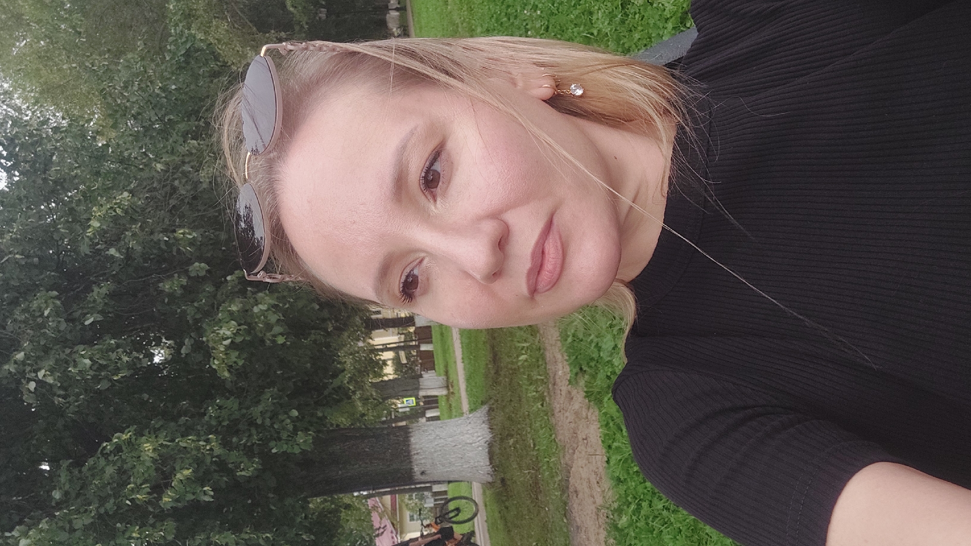 A woman with blonde hair, wearing sunglasses on her head and a black ribbed t-shirt, is shown in a close-up portrait outdoors. She is looking to her left with a neutral expression. The background includes green grass, trees, and a paved path. In the distance, a tree painted white at its base and a bicycle are visible. The scene appears to be in a park or public space in the city of Votkinsk, Russia.