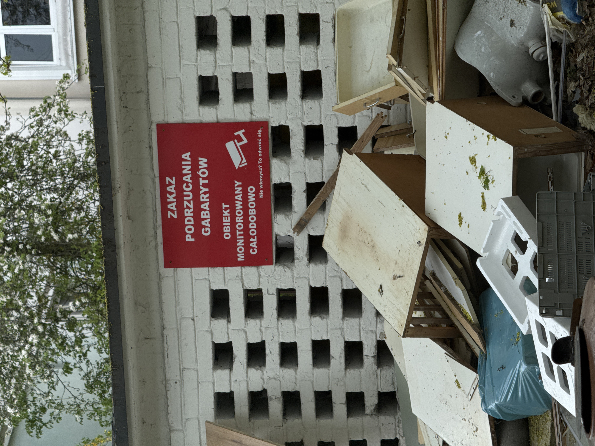 This image captures a scene of illegal waste dumping next to a building, likely in an urban or suburban area. The perspective is rotated 90 degrees clockwise.
On the right, a large pile of discarded items is visible. This includes various pieces of broken furniture like wooden cabinets and shelves, a white ceramic object resembling a sink or toilet base, white polystyrene packaging, a grey plastic crate, and a light blue or greenish trash bag. There is also general debris, dirt, and some green plant matter growing amidst the refuse.
To the left, a white brick wall forms part of a building, characterized by a grid-like pattern of square indentations or openings. Mounted on this wall is a prominent red rectangular sign with white Polish text. Further left, beyond the building, a tree or large bush with green leaves and white blossoms is visible, suggesting spring or early summer. Part of a building window can be seen through the foliage.
The red sign explicitly warns against illegal dumping. The text reads:
- "ZAKAZ PODRZUCANIA GABARYTÓW" (Prohibition of dumping bulky waste)
- "OBIEKT MONITOROWANY CAŁODOBOWO" (Object/Area monitored 24/7)
- Below this, smaller text states: "Nie wierzysz? To odwróć się." (Don't believe it? Turn around.), accompanied by an icon of a CCTV camera.
The scene depicts a clear act of environmental neglect or illegal disposal of bulky waste, with an official warning sign indicating that the area is under surveillance to deter such activities. The lighting suggests it is daytime, possibly overcast. Given the location context, this scene is set in Warsaw, Poland.
