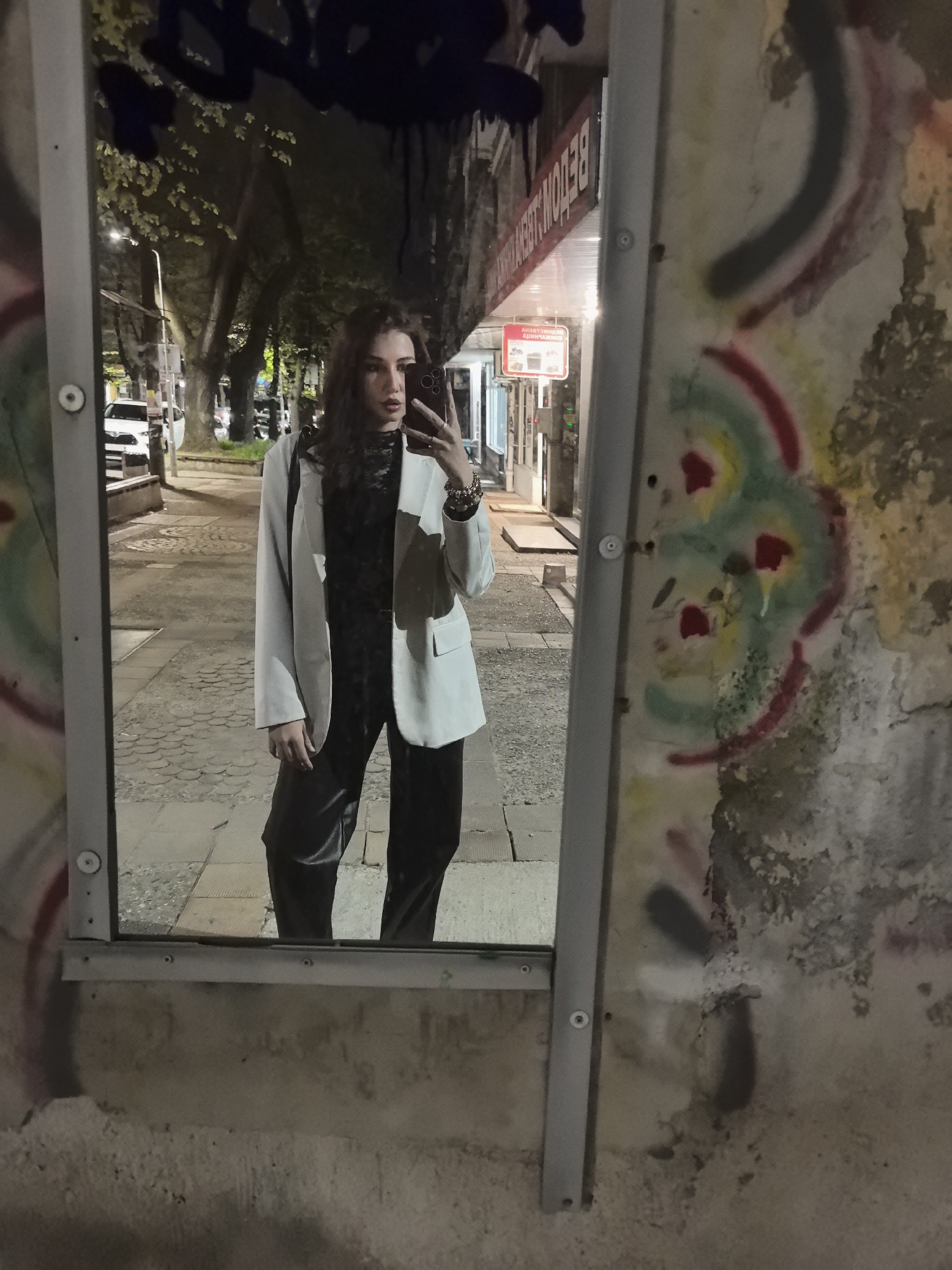 A young woman stands reflected in a tall, narrow mirror attached to a graffiti-covered wall on a city street. She is wearing a light-colored blazer over a black top and dark trousers. She holds a smartphone up to take a selfie. The background visible in the mirror shows a street with trees and buildings. A white car is visible parked on the left side of the street. A sign above a storefront in Cyrillic is also visible, indicating the location is likely in Bulgaria. Based on other cues, the location is likely Stara Zagora, Bulgaria.