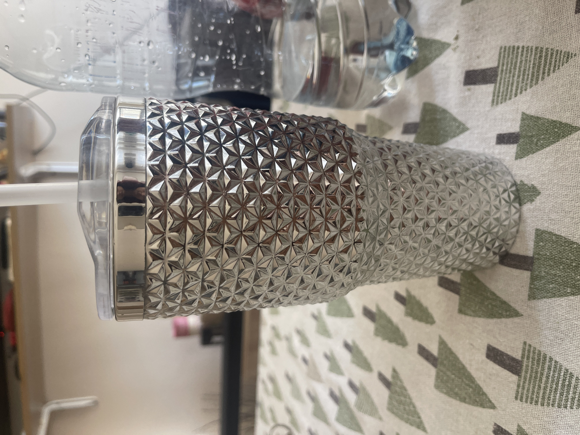 The image depicts a close-up shot of a silver, textured tumbler with a clear plastic lid and a white straw. The tumbler has a raised geometric pattern. To the right of the tumbler is a partially visible plastic bottle containing a clear liquid with blue accents and condensation on its surface. Both the tumbler and bottle are placed on a tablecloth with a repeating pattern of stylized green and grey trees. In the background, out of focus, are shelves with various objects, including what appear to be food items. The lighting suggests an indoor setting, possibly a home or a cafe. The image does not provide any specific landmarks or clues to pinpoint a location within Varna, Bulgaria, or any city. There are no people or discernible actions taking place.