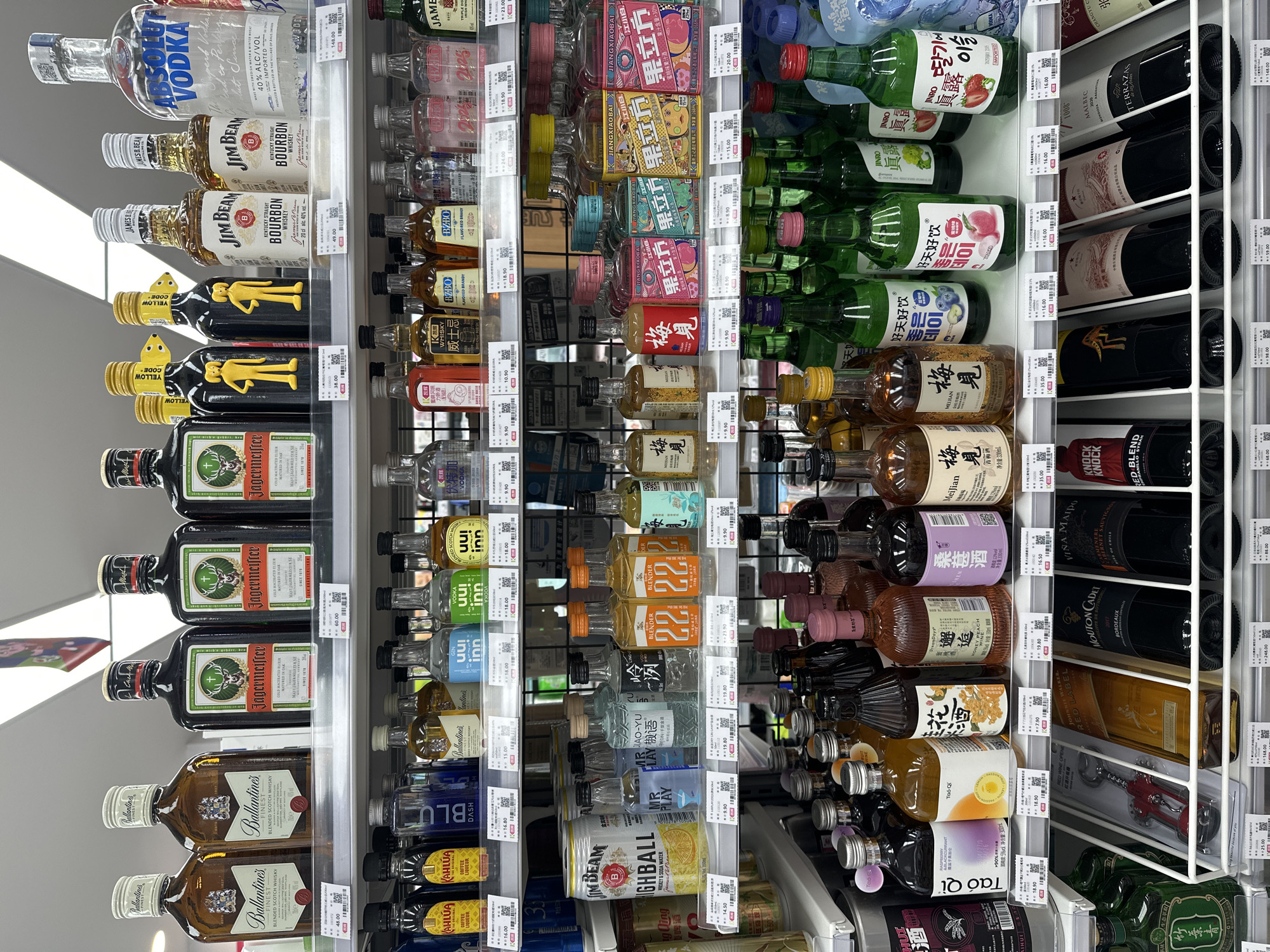 The image shows a well-stocked liquor store shelf in the city of Chongqing High-tech Zone, China. The shelves are filled with various bottles of alcoholic beverages, predominantly spirits and wine, arranged in an organized manner. The lighting is bright and even, typical of a retail environment. No people are visible in the frame.
On the top shelf, several well-known brands of whisky and vodka are displayed, including Ballantine's, Jägermeister, Jim Beam, and Absolut Vodka. These bottles vary in size and color, creating a visually diverse display. The prices are clearly marked on small white labels placed below each product.
The middle shelf features a mix of smaller spirit bottles, likely miniatures or sample sizes, alongside some larger bottles. Brands such as Kahlua, Blue Sapphire, and various Chinese liquors are present. Some bottles have colorful labels featuring images of fruits or traditional designs. There are also some large cans or bottles of what appears to be a beverage mix.
The bottom shelf showcases a variety of wine bottles, predominantly red wine, from different regions. Brands like Mouton Cadet, Vina Maipo, and Terranaz are visible, along with some less identifiable bottles. The wines are displayed upright in a neat row, with their labels facing forward. Some bottles are also placed on their sides in display racks. Additionally, there are some smaller bottles of what appear to be traditional Chinese liquor or flavored spirits on the lower portion of this shelf.
The overall environment suggests a retail setting designed for showcasing and selling alcoholic beverages. The presence of price tags indicates an active commercial transaction environment. The display is orderly, with products neatly arranged on illuminated shelves. The background shows the continuation of the shelving units and the store's ceiling structure, implying a typical supermarket or liquor store aisle.