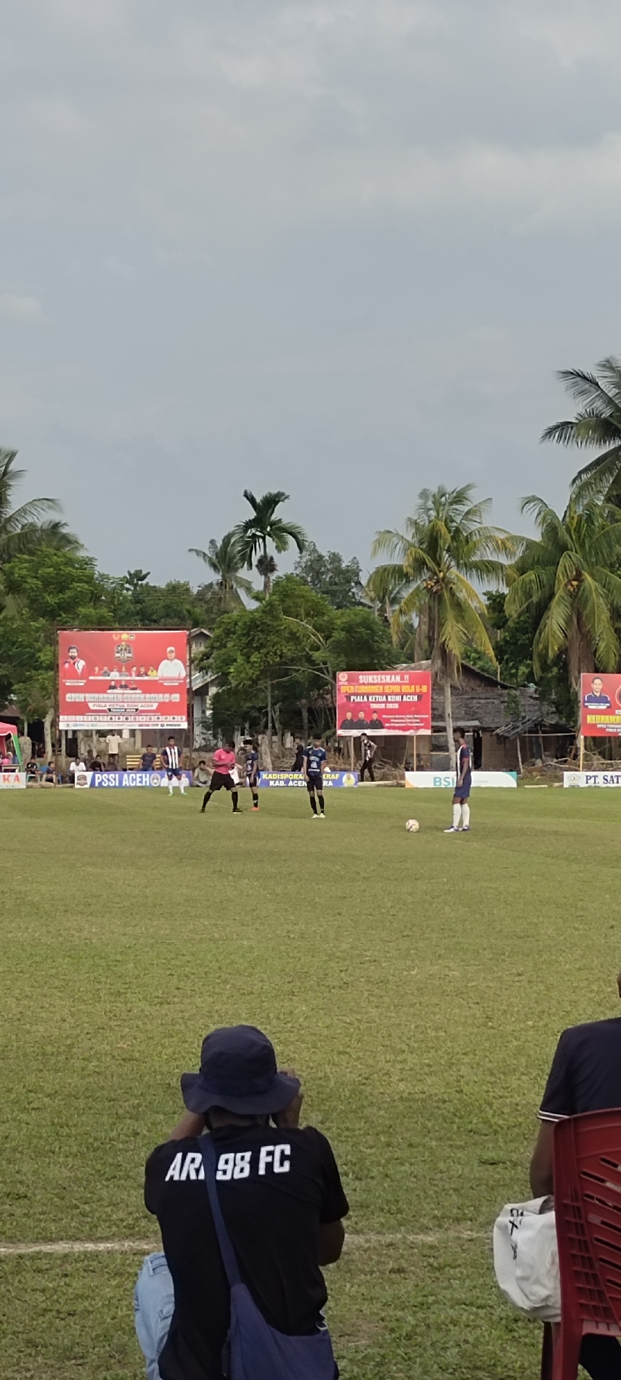 The image captures a football match in progress at a field in Kilometer Delapan, Indonesia. In the foreground, the back of a person wearing a black t-shirt with "AR 98 FC" printed on it is visible. They are wearing a dark blue bucket hat and are holding what appears to be a camera or phone. To their right, another person is seated on a red chair.
The middle ground features a grassy football field with several players in uniform. One player in a blue and white striped jersey is standing near a football, while another player in a blue jersey and another in a black and white striped uniform are also present. A referee in a pink shirt is observing the game. In the background, banners with text are displayed, and palm trees and structures are visible. The sky is overcast.
