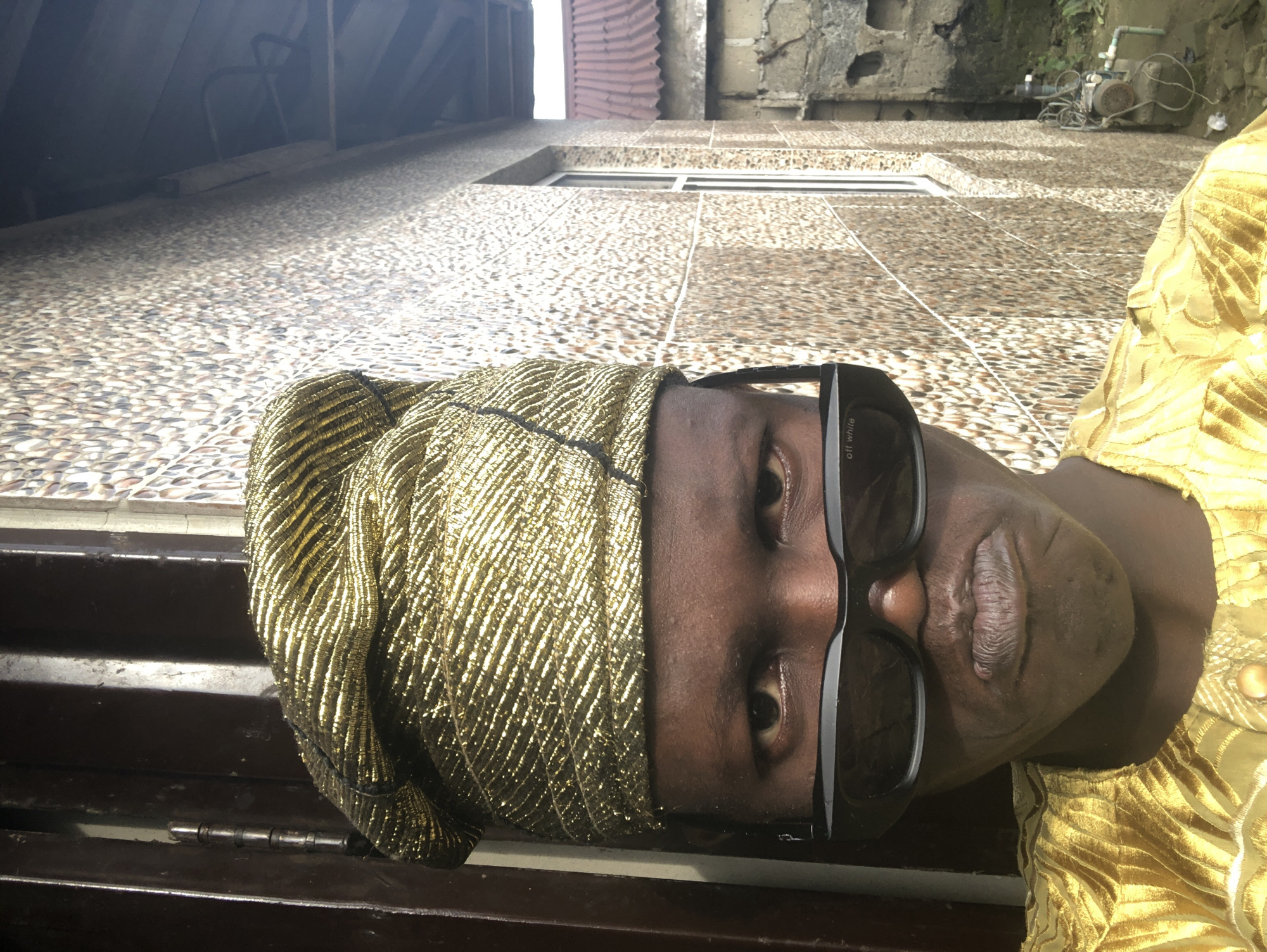 The image is a medium shot of a person from the chest up, facing the camera directly. They are wearing a golden, textured headwrap and a matching golden, patterned shirt. Large, black sunglasses with white lettering on the left lens are worn, obscuring the upper part of their face. The person's lips are pursed, creating a distinctive expression. The background consists of a textured wall with a pattern of small, multi-colored tiles on the right, and a dark, vertical surface on the left, possibly a door or window frame. The lighting suggests an outdoor setting, with shadows indicating sunlight. There is a hint of a building's roofline and some foliage in the upper right corner, suggesting an urban or semi-urban environment. The overall impression is that of a posed portrait. The context of Ado, Nigeria, suggests this is likely a residential or commercial area.