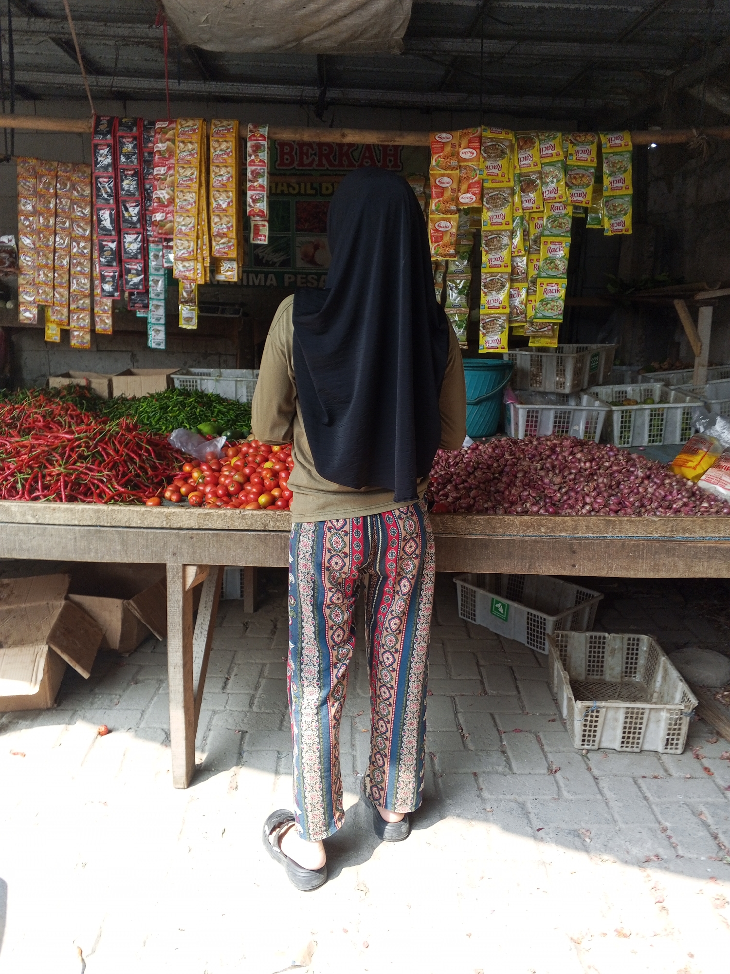 A person, viewed from behind, stands at an outdoor market stall in Ciruas, Indonesia. The individual wears a dark hijab, a tan long-sleeved shirt, and colorful patterned pants. They are facing a table laden with produce, including large piles of red chili peppers, green chilies, tomatoes, and red onions. Various packaged goods, such as spices, hang from a wooden structure above the stall. Several plastic crates and cardboard boxes are visible around the stall, some empty and others containing produce. The ground is paved with grey bricks, and sunlight casts shadows across the scene, indicating it is daytime.